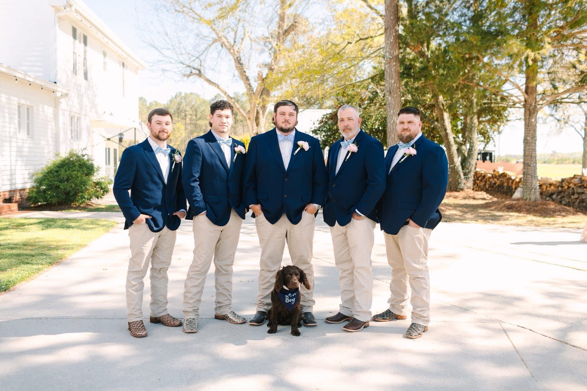 Groom and four groomsmen in matching navy blazers and khaki pants pose on the sunlit driveway at Scarborough House with a chocolate spaniel wearing a ring bearer bandana in front.