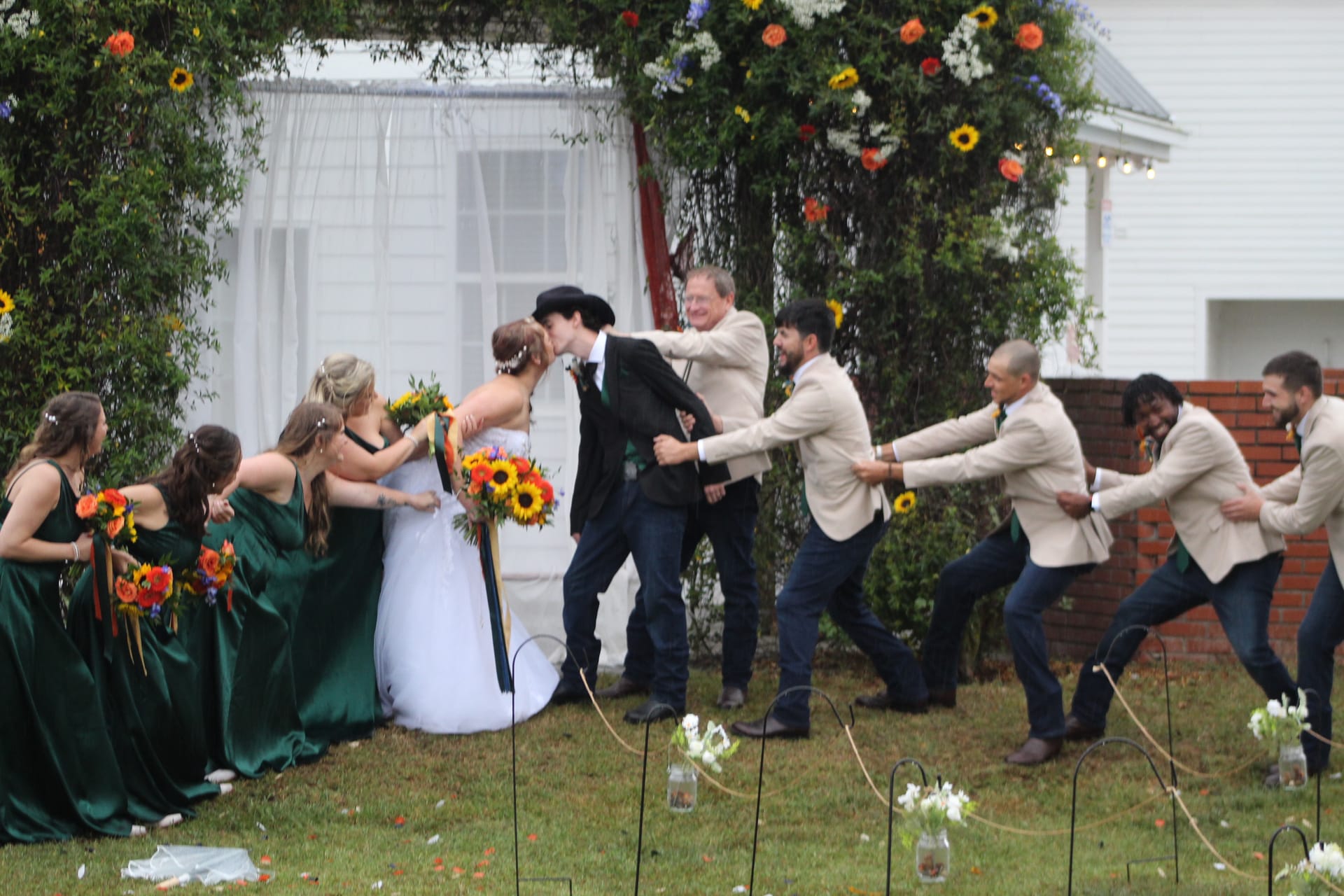 Bride and groom share their first kiss under a floral arch while bridesmaids and groomsmen playfully pull them apart on the ceremony lawn at Scarborough House.