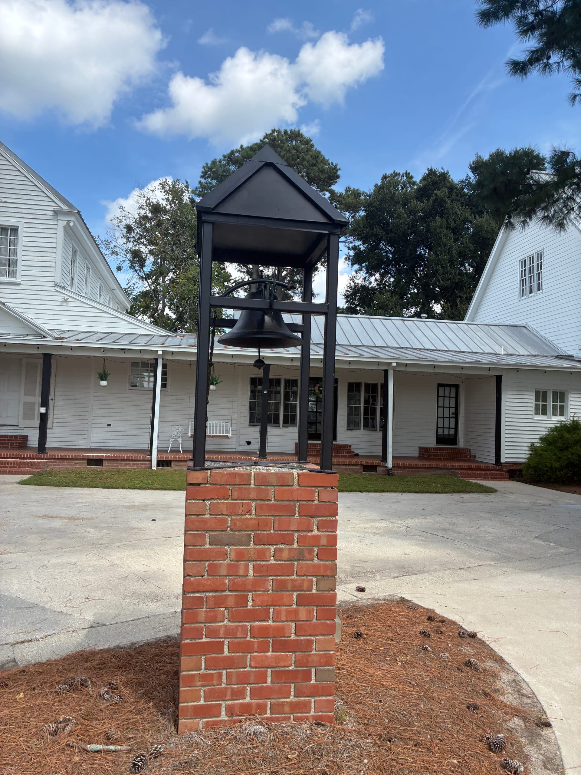 Black iron bell mounted on a brick pedestal in front of the white historic Scarborough House manor with covered porch and pine trees under a partly cloudy sky.
