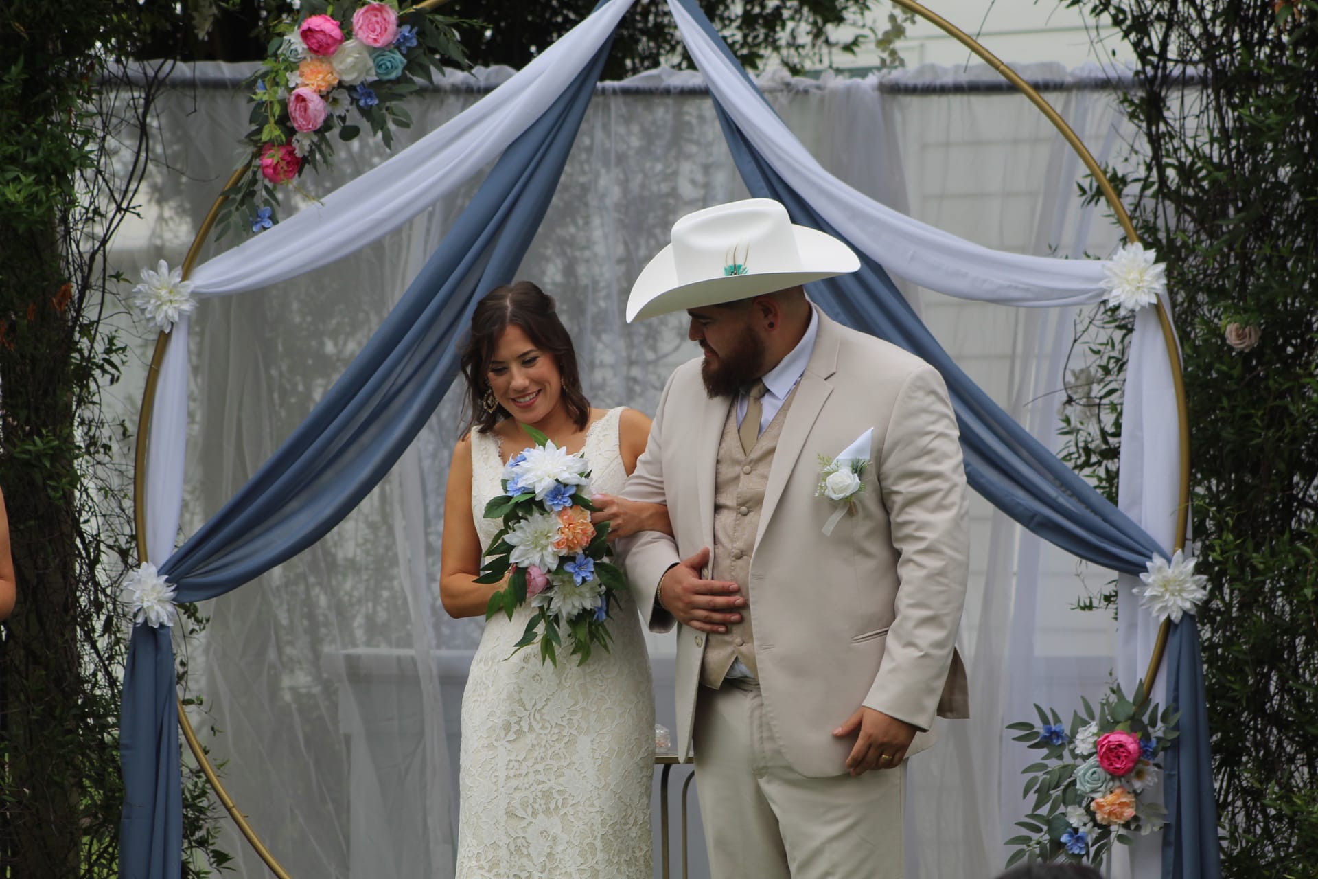 Smiling bride in lace gown holding a colorful bouquet stands with her groom in a tan suit and white cowboy hat beneath a draped circular arch decorated with pink and blue flowers at an outdoor wedding ceremony.