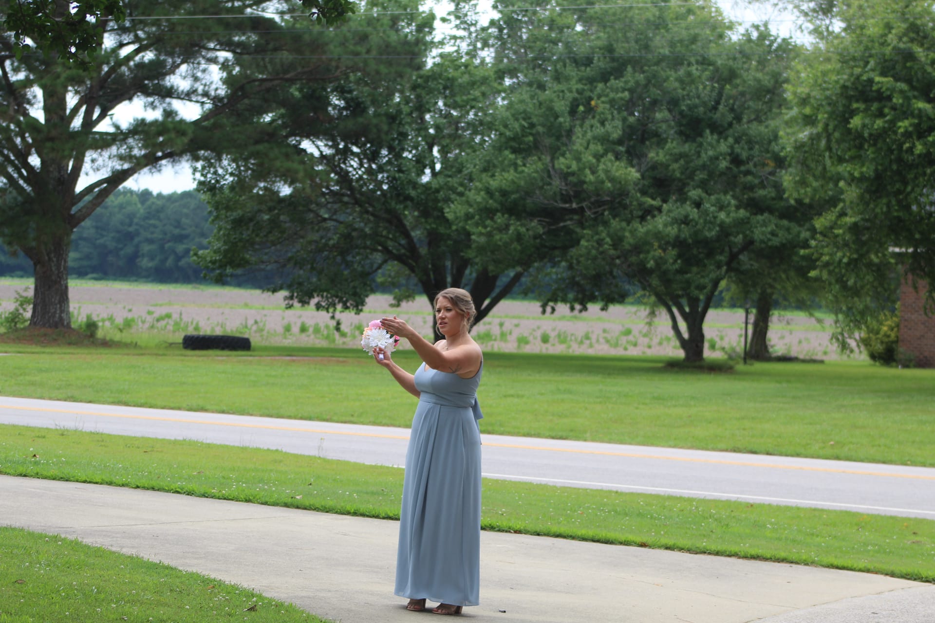 A bridesmaid in a flowing dusty blue gown holds a white and pink bouquet while standing on a sidewalk with green lawns and open farmland fields in the background at Scarborough House.
