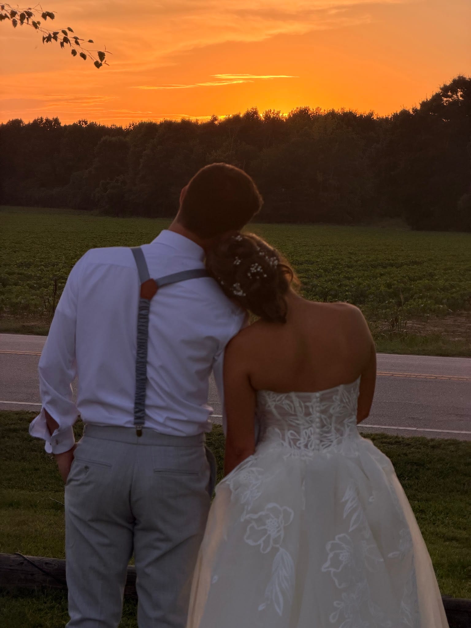Bride and groom seen from behind with heads together watching a glowing orange sunset over open farmland fields at Scarborough House estate.