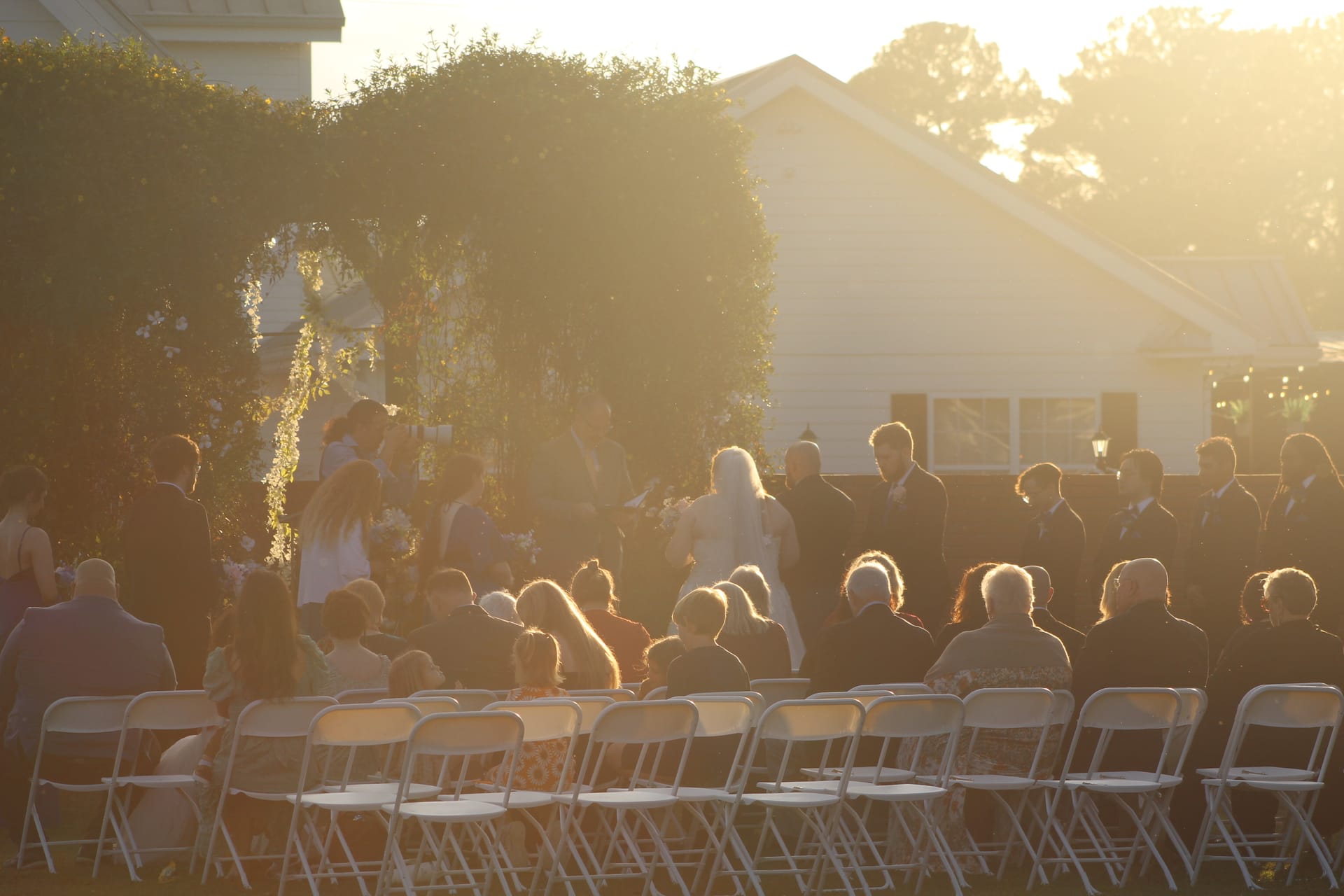 Guests seated in white folding chairs watch a wedding ceremony bathed in warm golden sunset light on the ceremony lawn at Scarborough House historic estate.