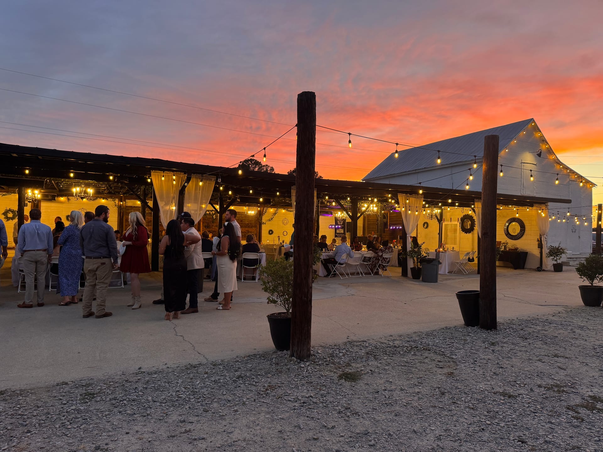 Guests mingle under warm string lights at an outdoor evening wedding reception beside a white barn venue beneath a dramatic orange and pink sunset sky.
