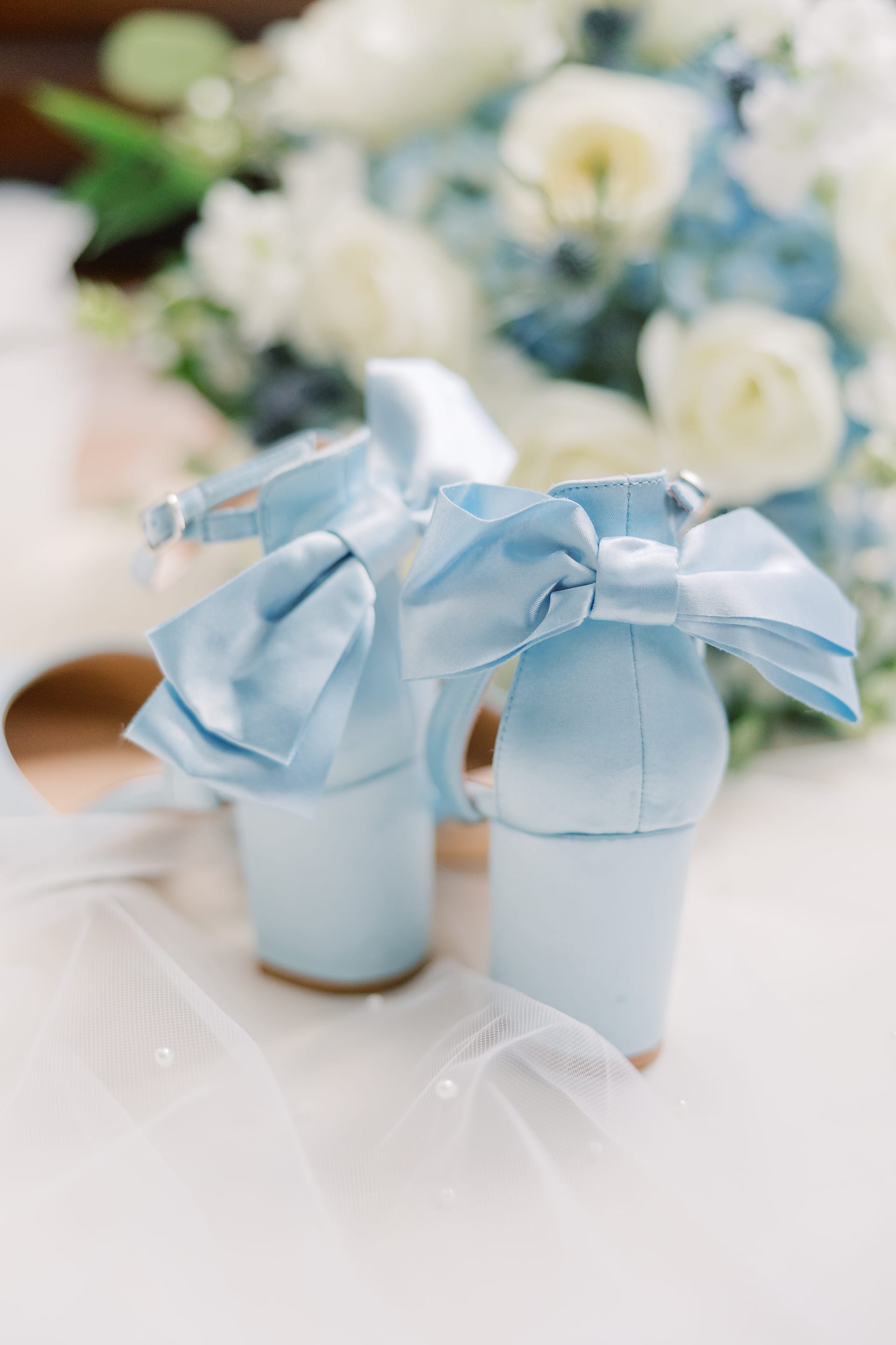 Pale blue satin block-heel wedding shoes with a large bow rest on a white veil beside a soft-focus bouquet of white roses and blue hydrangeas.