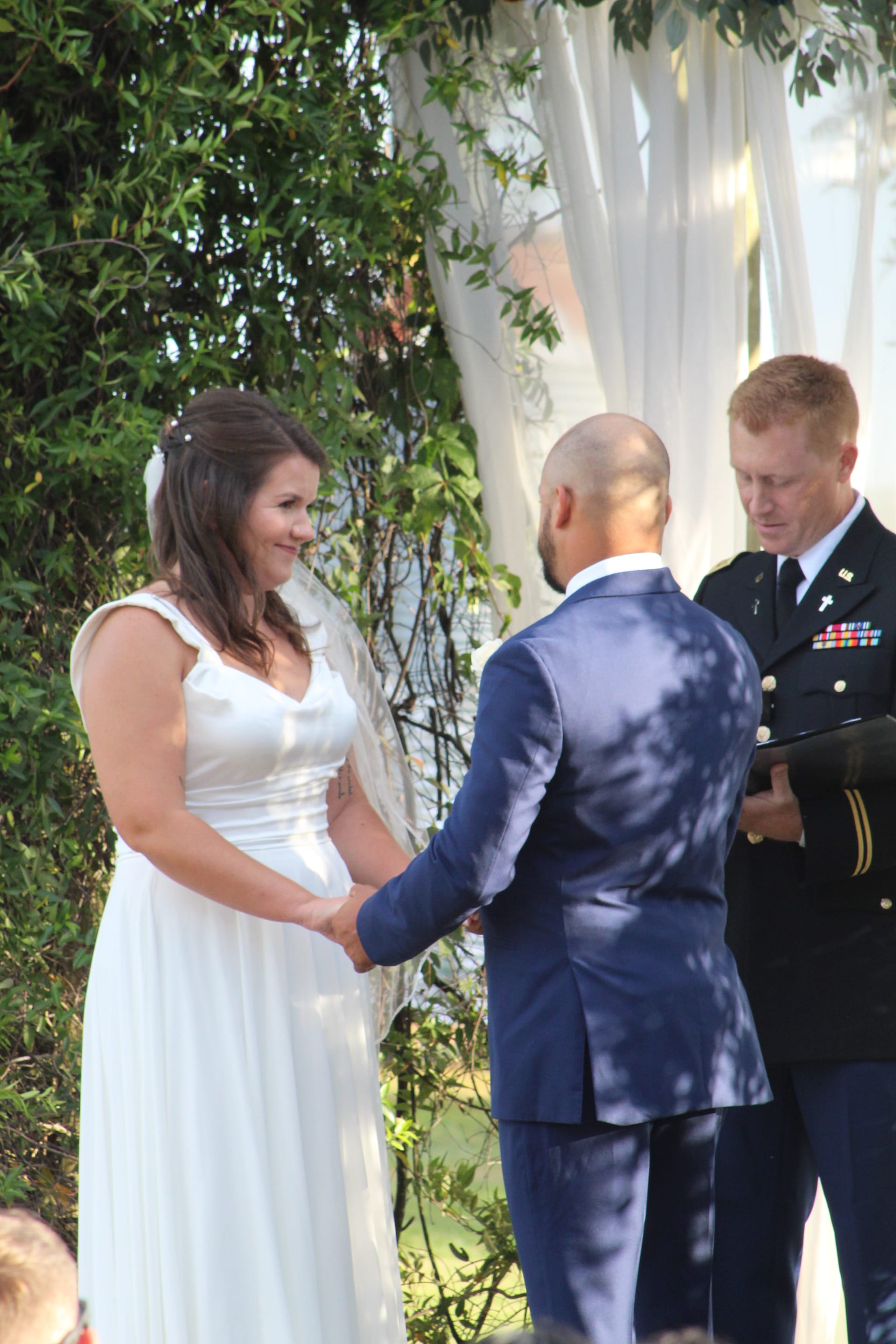 Bride in a flowing white gown smiles warmly as she exchanges vows with her groom while a military officer officiates the outdoor wedding ceremony amid lush greenery and draped white fabric.