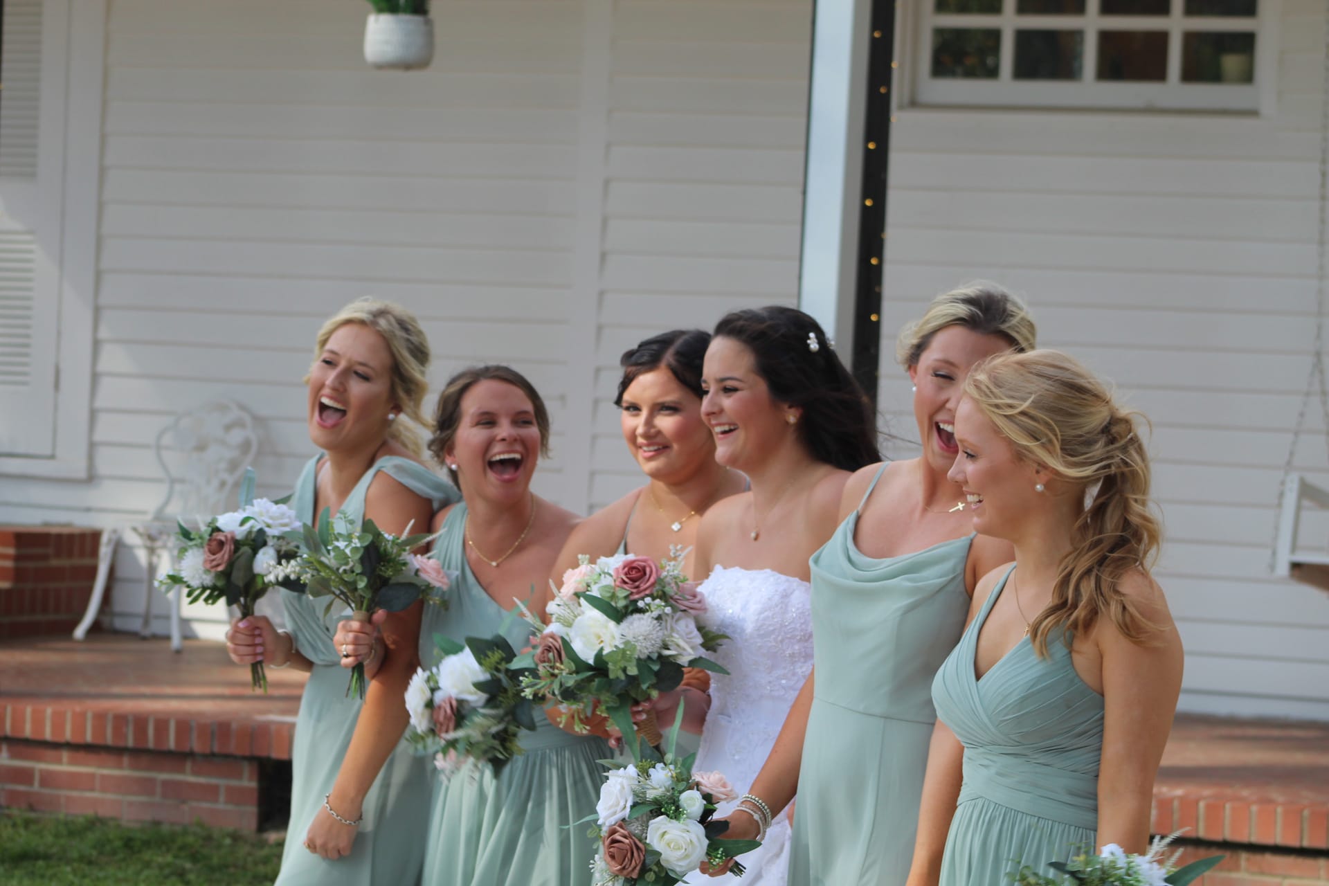A laughing bride in white lace and five bridesmaids in sage green gowns holding floral bouquets stand together in front of the historic white manor house at Scarborough House.