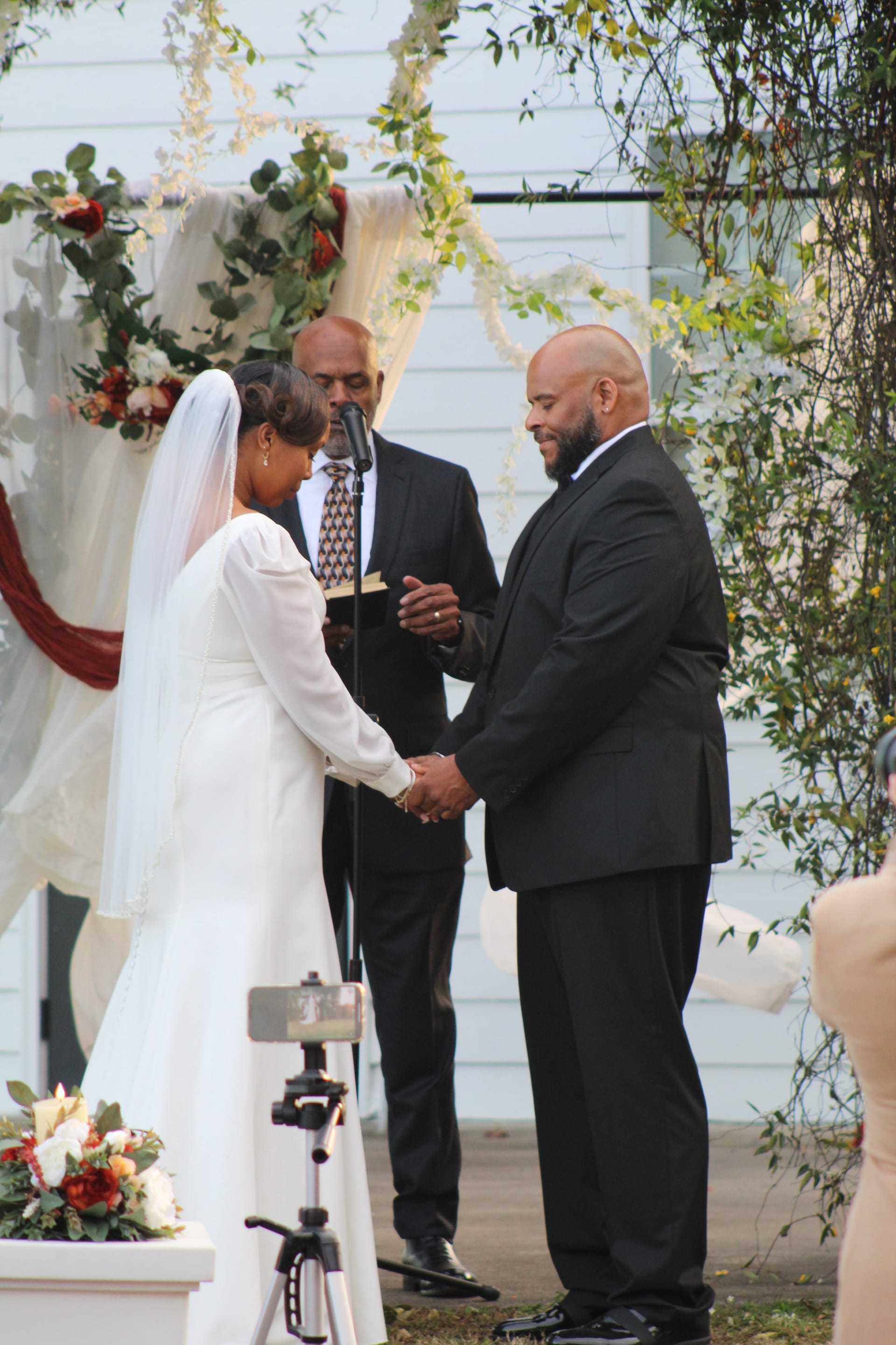 Bride in white gown and veil holds hands with groom in black suit during outdoor wedding ceremony beneath a floral arch at a historic estate venue.