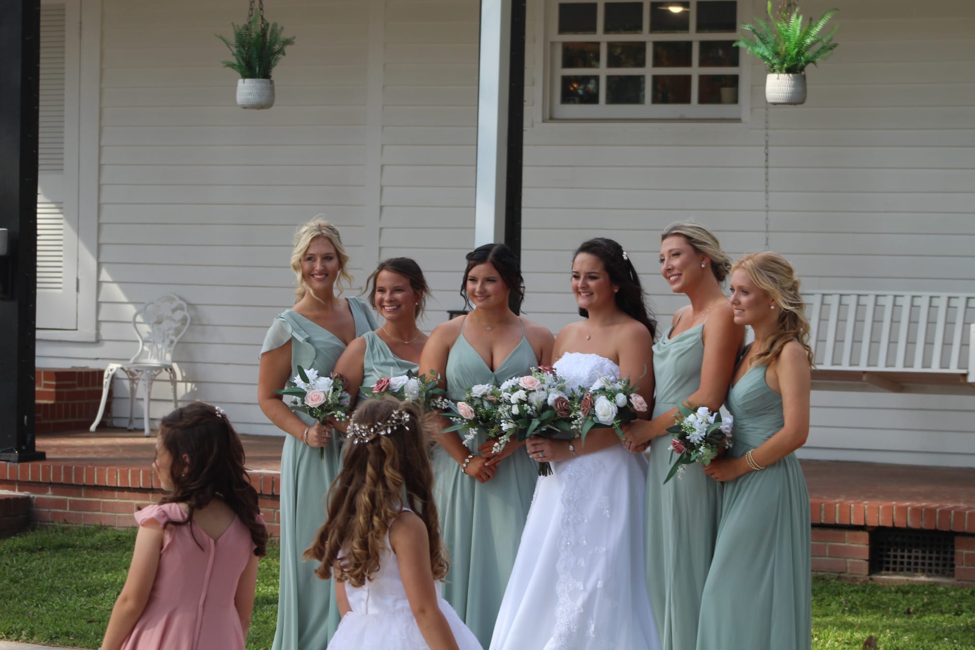 Bride in white gown surrounded by bridesmaids in sage green dresses holding floral bouquets with flower girls in front of the historic Scarborough House manor.
