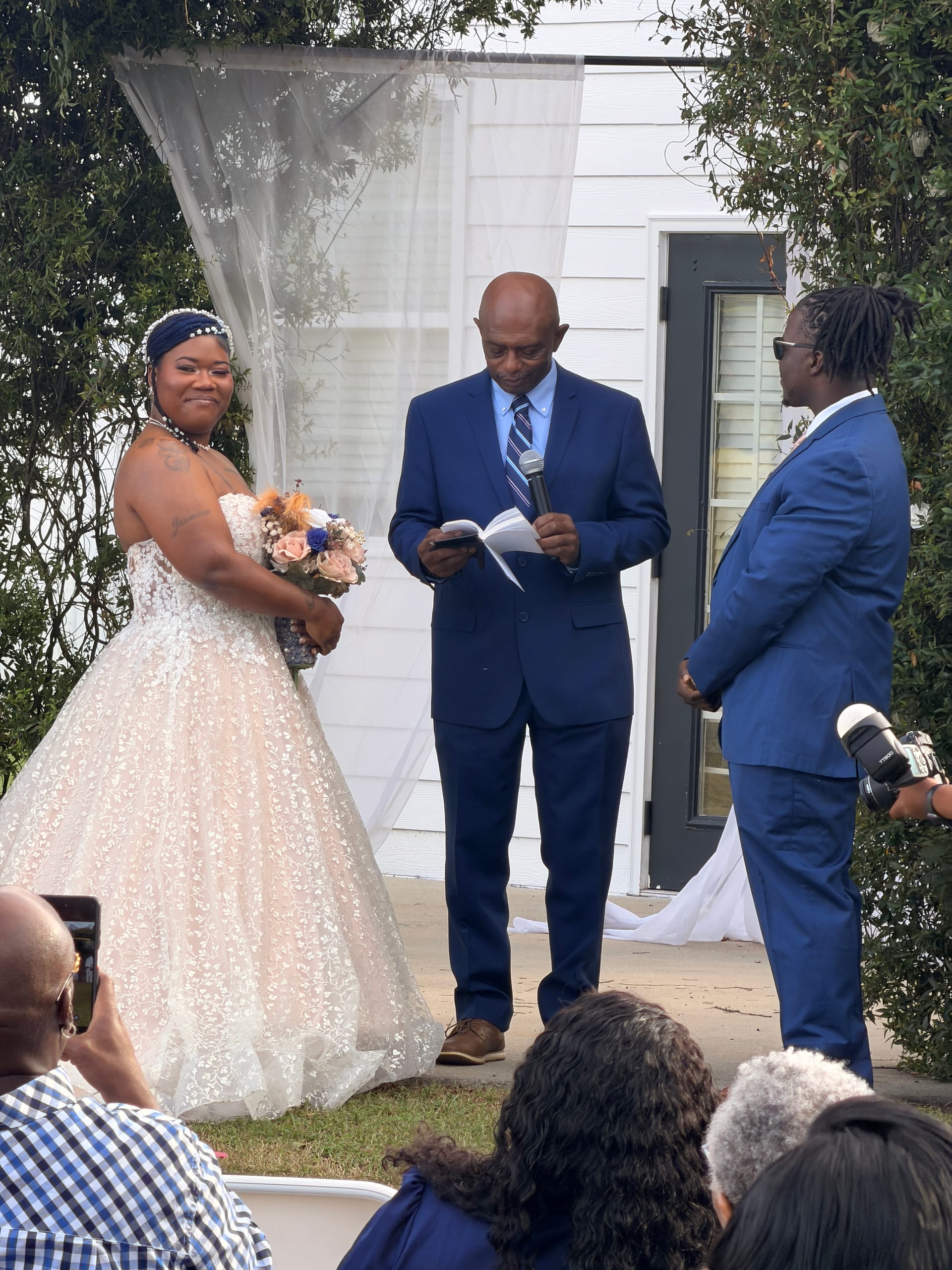 A bride in a white ballgown holding a floral bouquet smiles during an outdoor wedding ceremony at Scarborough House as the officiant reads from a book beside the groom in a navy suit.