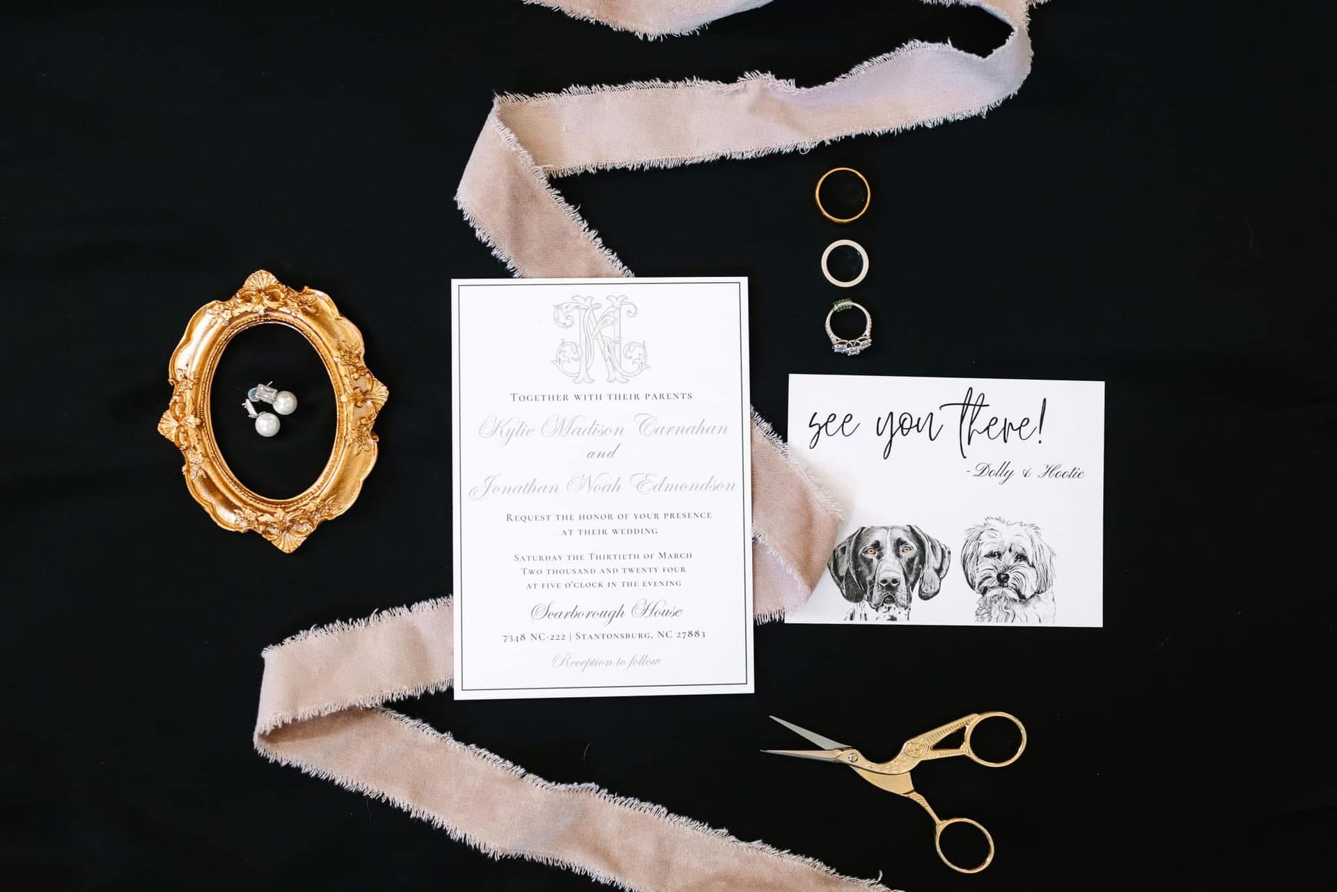 Elegant wedding invitation flatlay for Kylie Madison Carnahan and Jonathan Noah Edmondson featuring calligraphy stationery, gold rings, pearl earrings in an ornate gold frame, silk ribbon, and a whimsical see-you-there card illustrated with two dogs named Dolly and Hootie at Scarborough House.