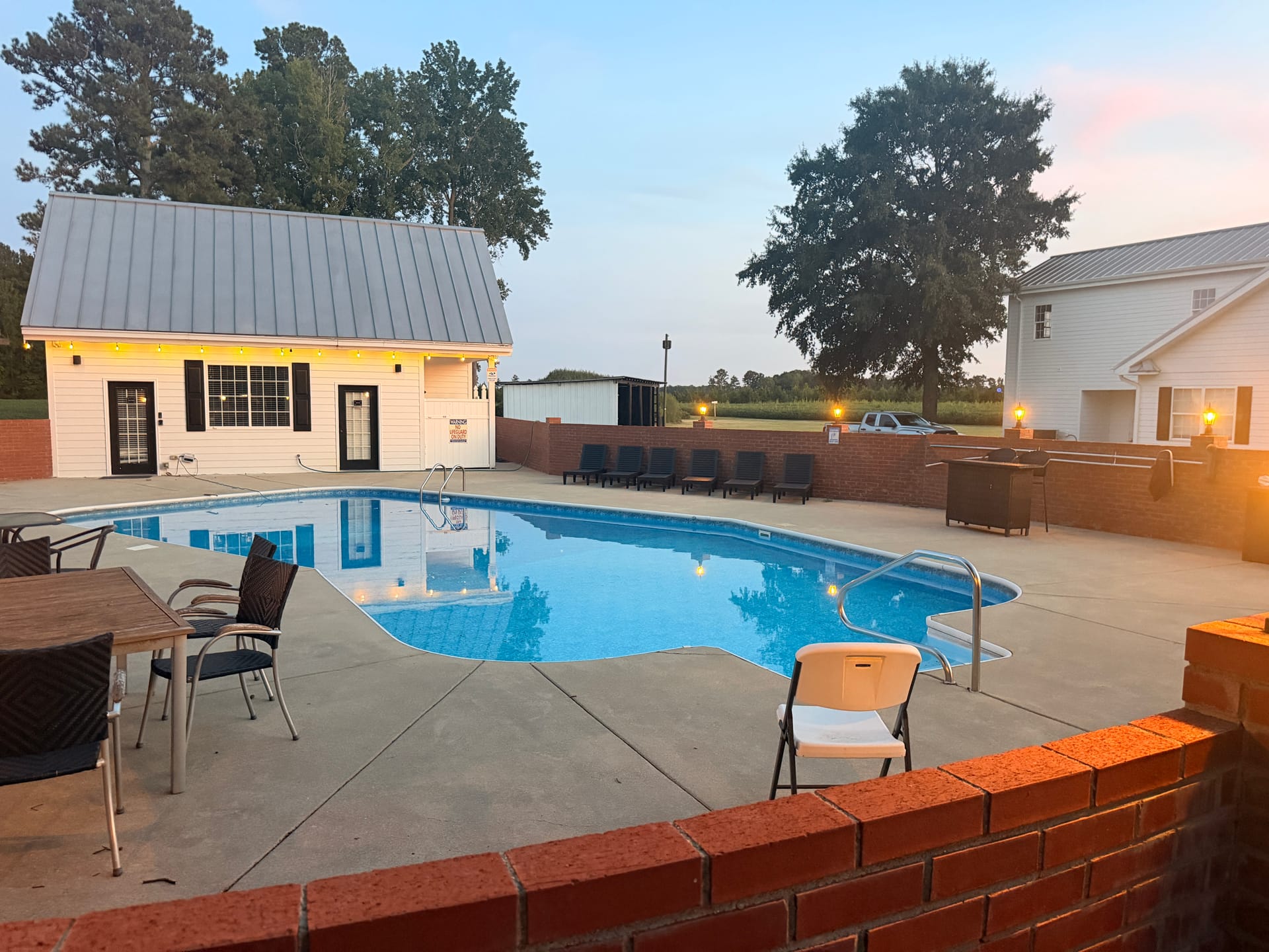 Twilight view of the Scarborough House outdoor pool surrounded by a brick patio, lounge seating, and a charming white pool house with string lights glowing at dusk.