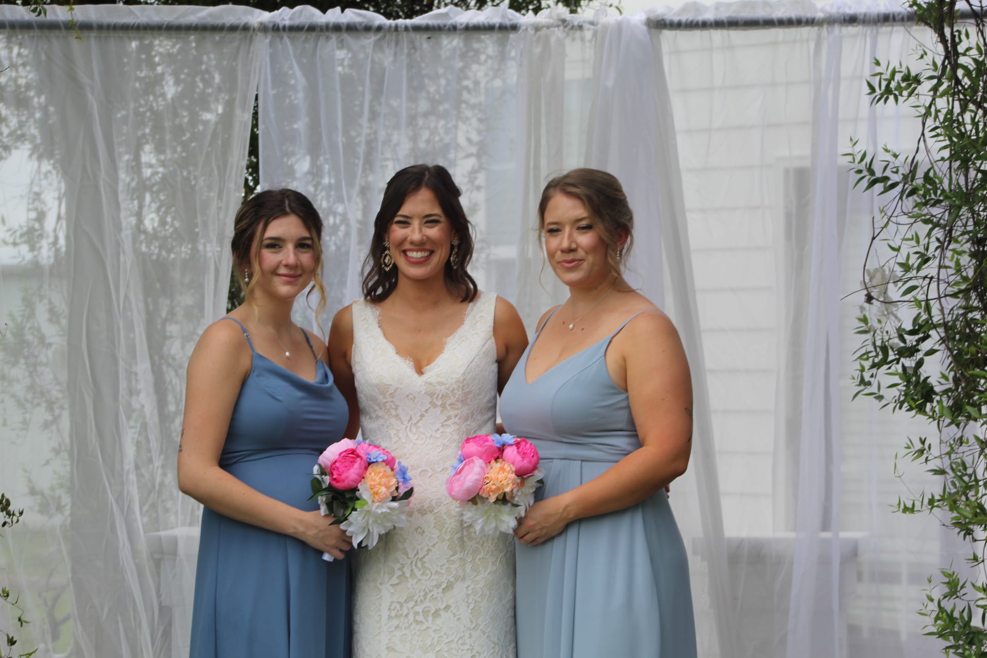 Bride in white lace gown smiles between two bridesmaids in dusty blue dresses holding colorful pink and peach bouquets beneath draped white fabric at Scarborough House.