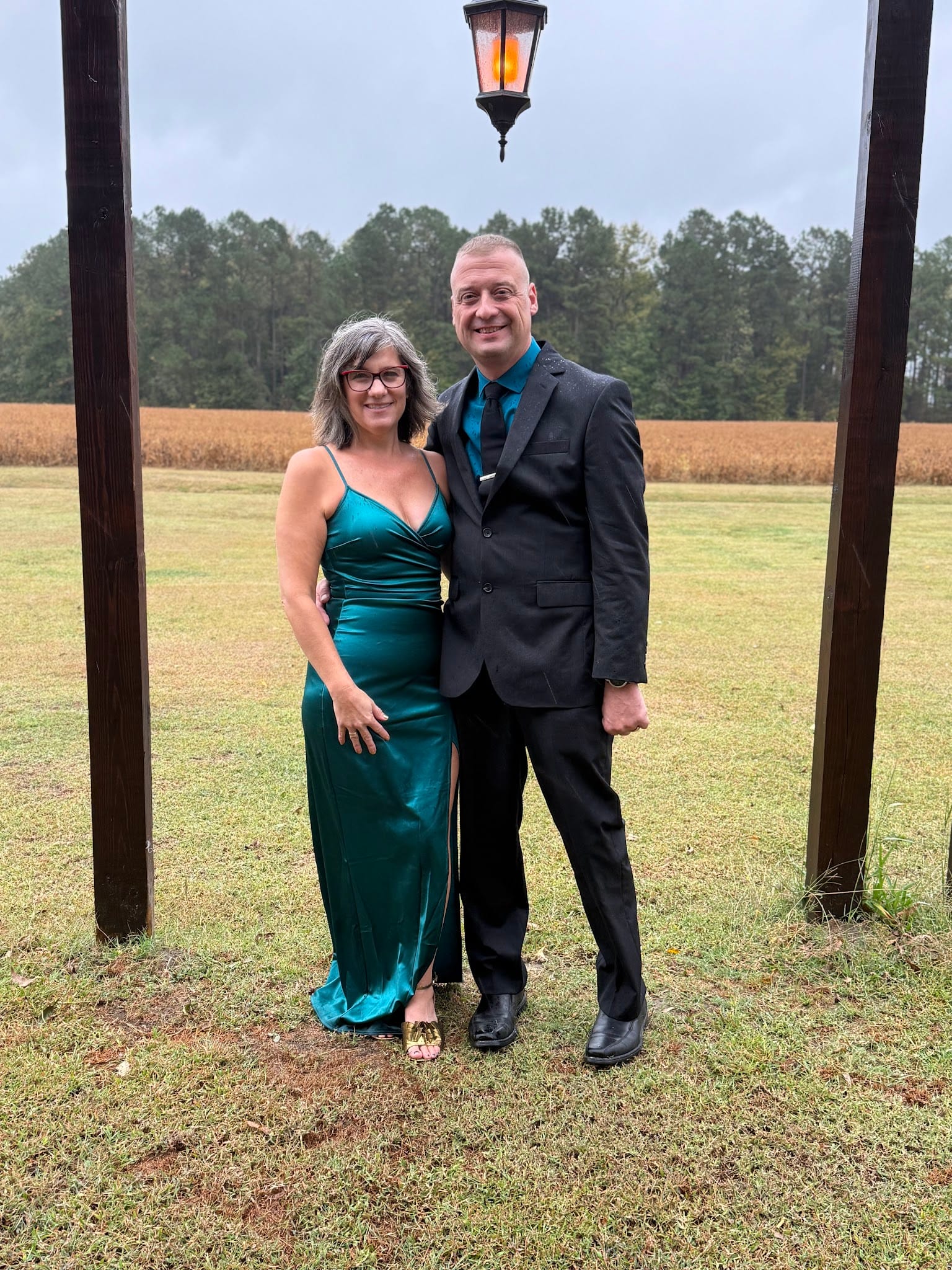 A smiling couple in formal attire — the woman in an emerald satin gown and the man in a charcoal suit with teal tie — pose between rustic wooden posts with a hanging lantern against open farmland fields at Scarborough House.