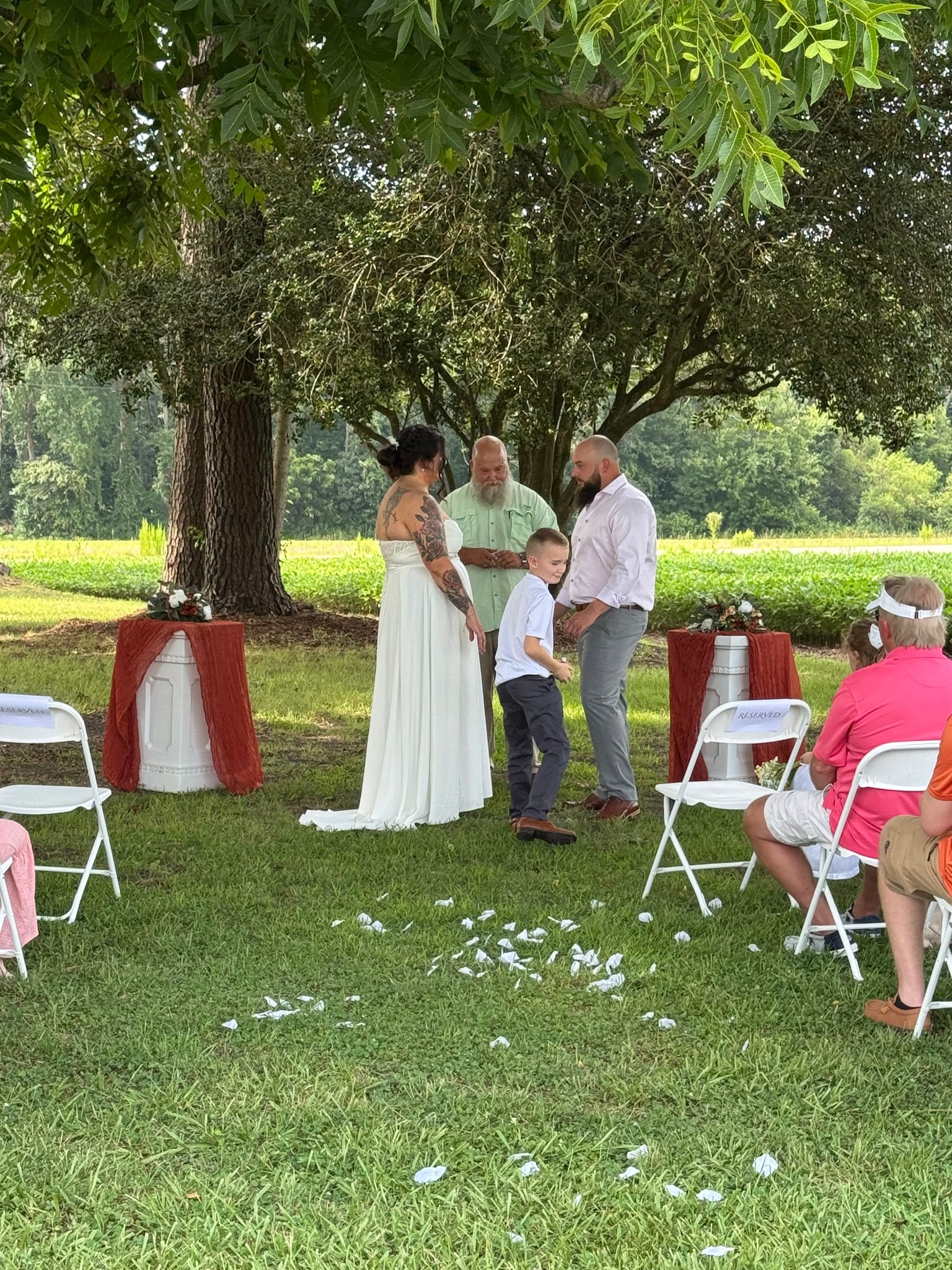 Bride in white gown and tattooed groom exchange vows beneath a large oak tree on the ceremony lawn at Scarborough House with a young child joining them at the altar.