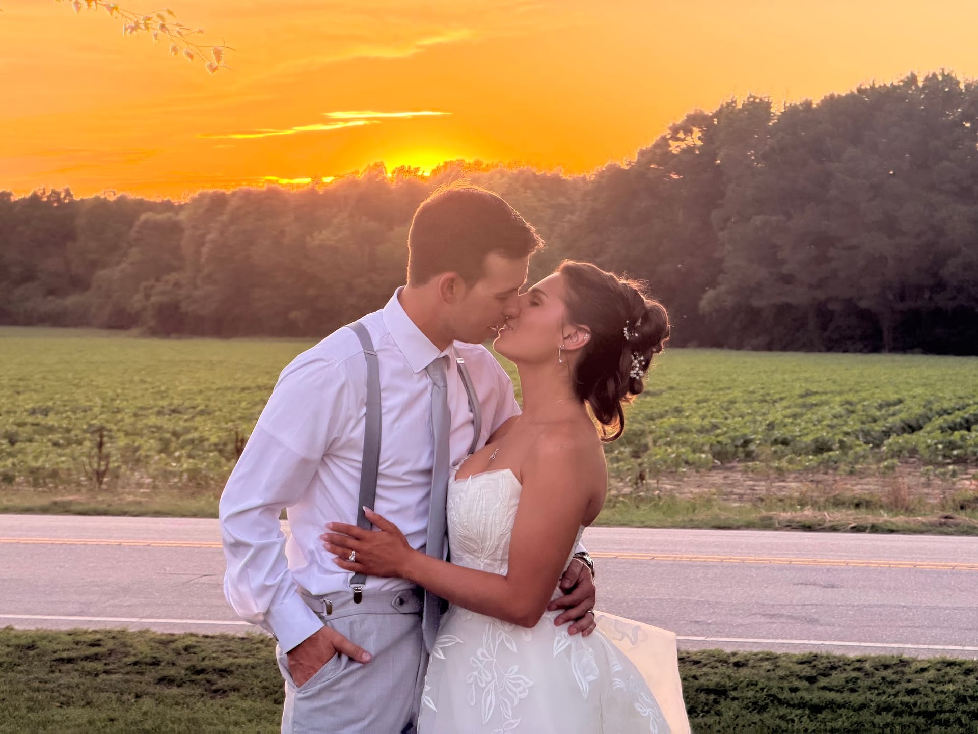 Bride and groom share a romantic kiss at golden hour with a vibrant orange sunset and green farmland fields behind them at Scarborough House.