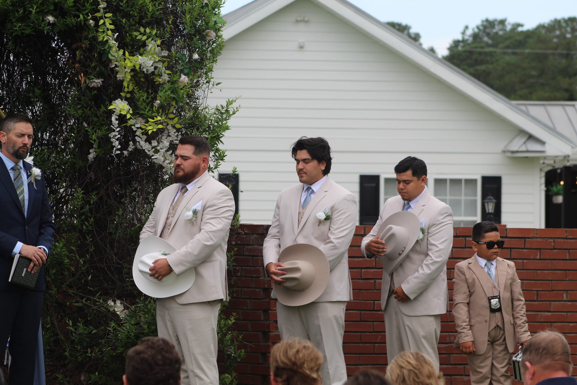 Three groomsmen in tan suits with boutonnieres hold cowboy hats solemnly during an outdoor wedding ceremony at a historic white estate venue.