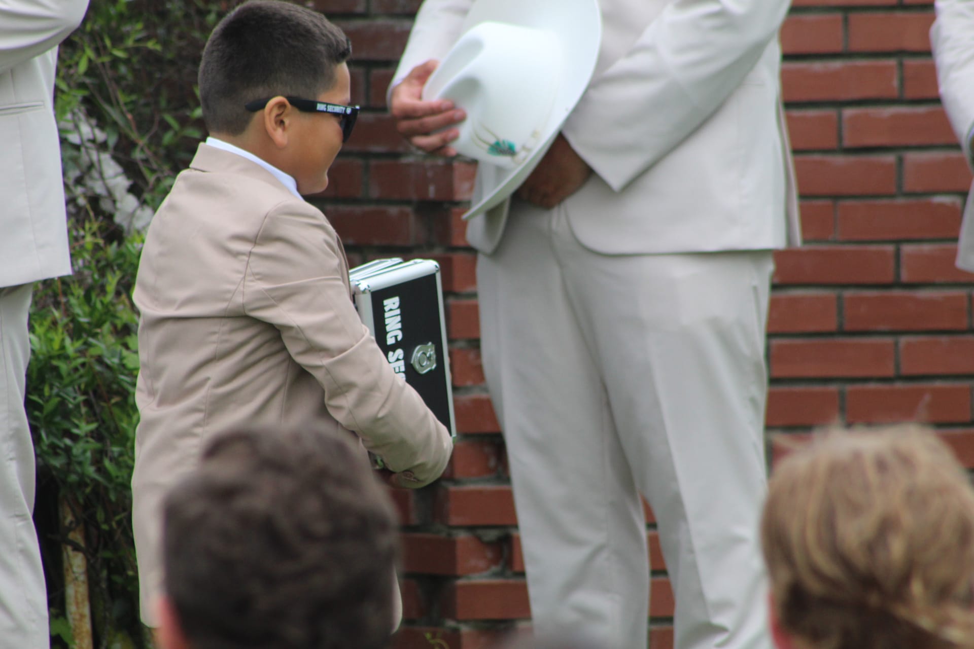 Young ring bearer in a tan suit and Ring Security sunglasses holds a black briefcase labeled Ring Security during an outdoor wedding ceremony.