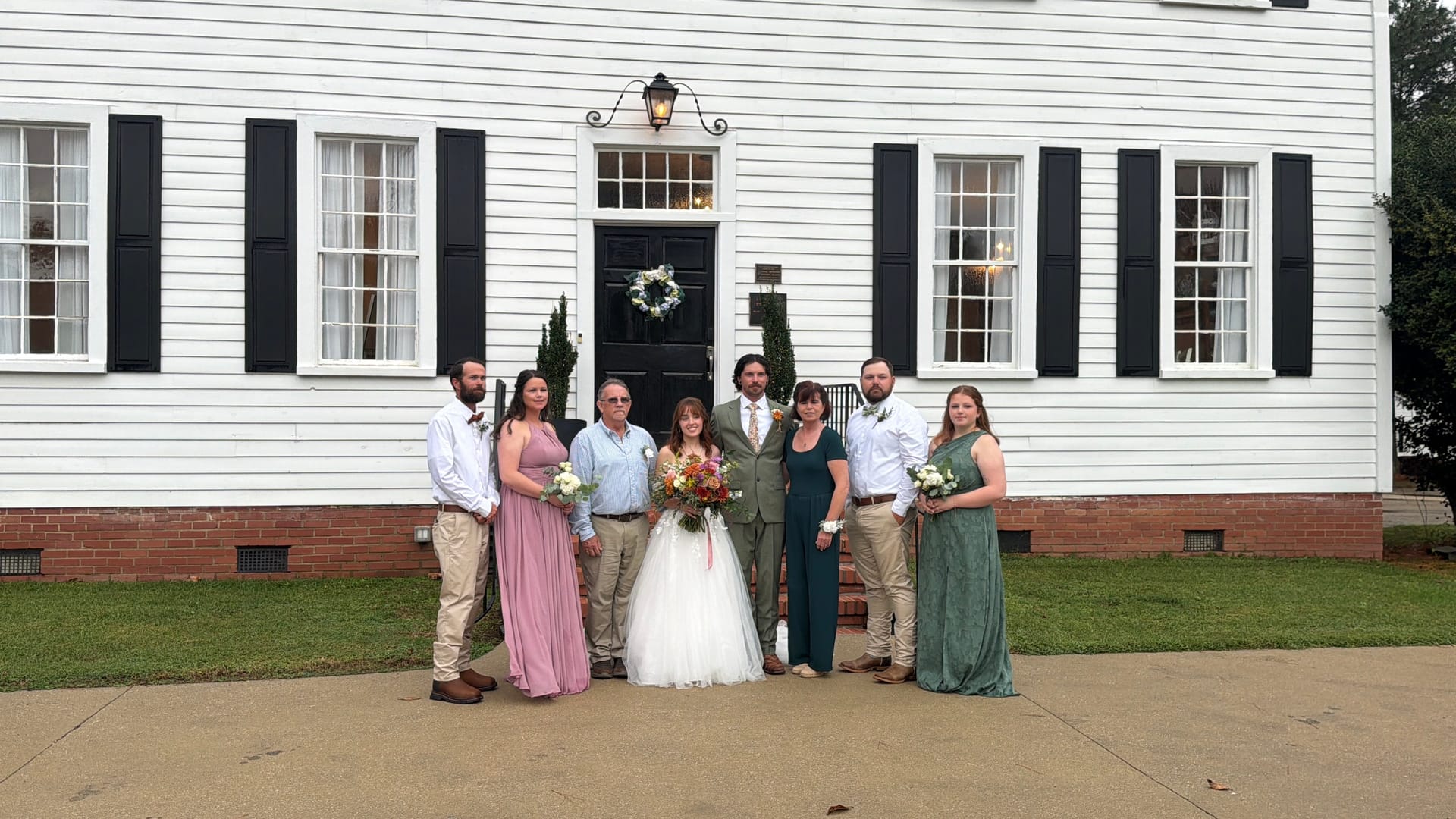 Wedding party and family members pose in coordinated attire in front of the white 1821 Scarborough House manor with black shutters and wreath-adorned door.