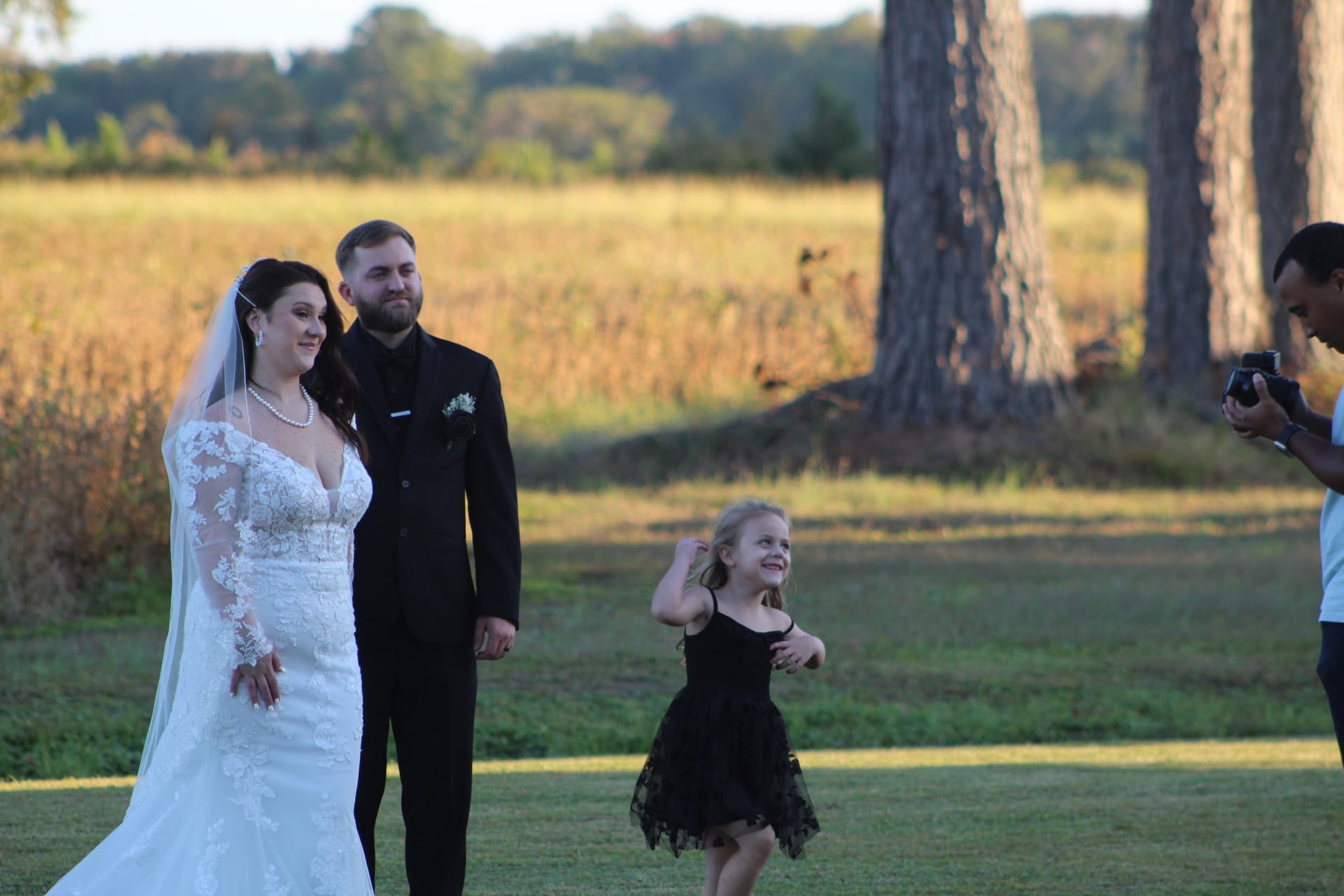 Bride in lace gown and veiled groom smile warmly as a young girl in a black dress twirls joyfully before them on the sun-drenched ceremony lawn at Scarborough House with golden farmland fields in the background.