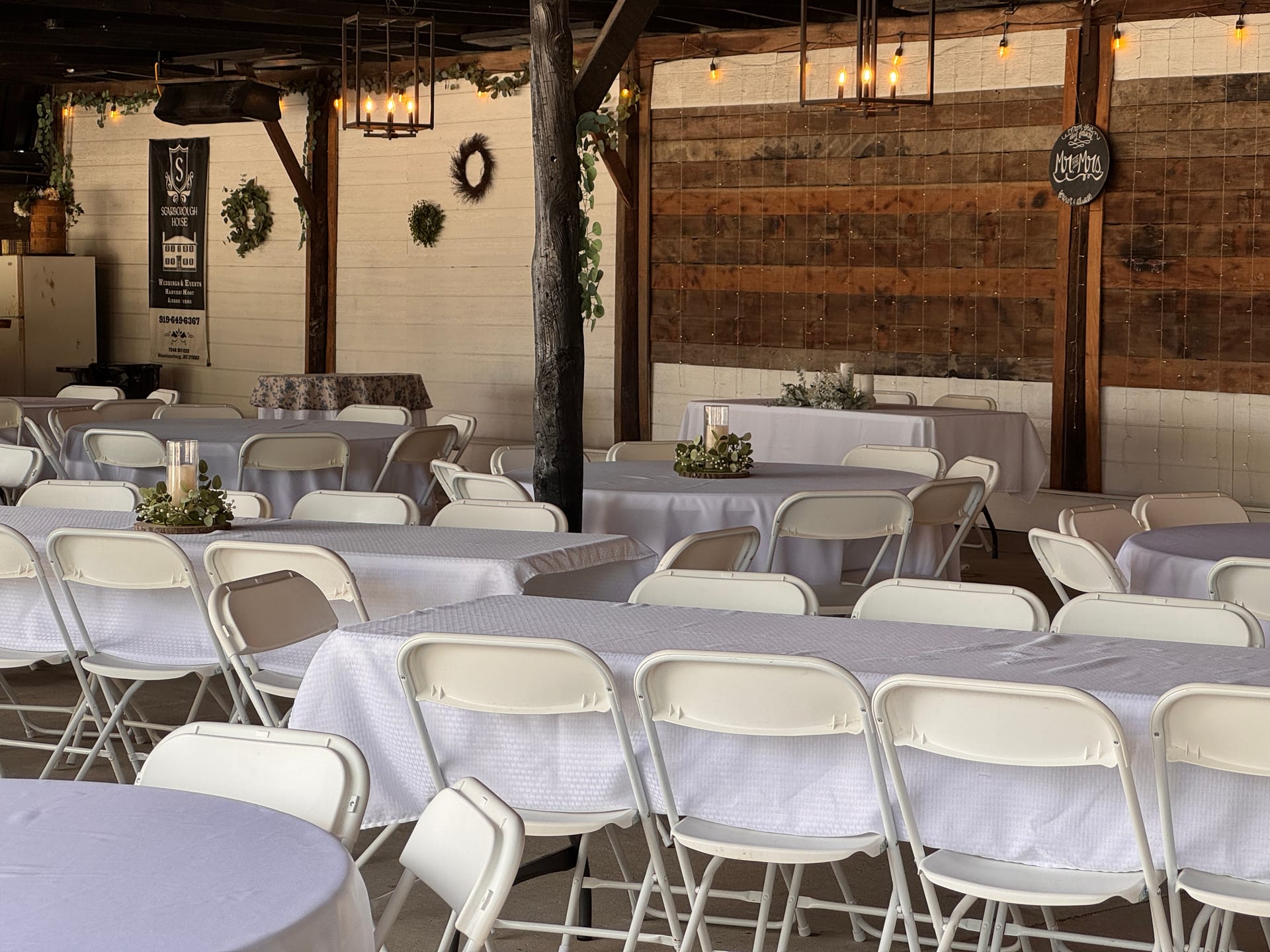 Rustic-elegant reception hall at Scarborough House set with long white-linen banquet tables, folding chairs, greenery centerpieces, wreath wall decor, and warm pendant lighting.