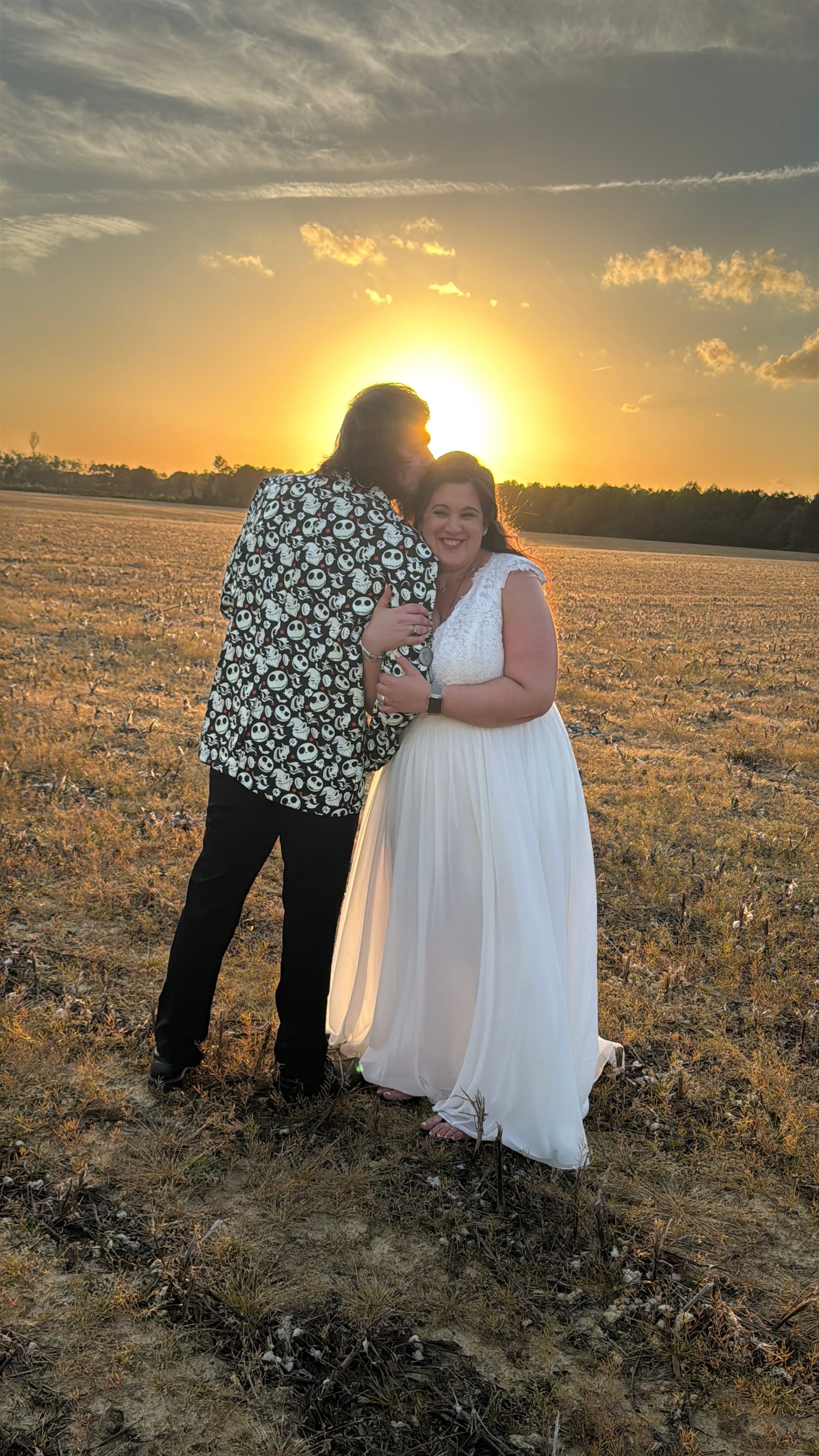 Bride in a flowing white gown smiles warmly as her groom embraces her in an open farmland field at Scarborough House, bathed in a glowing golden sunset.