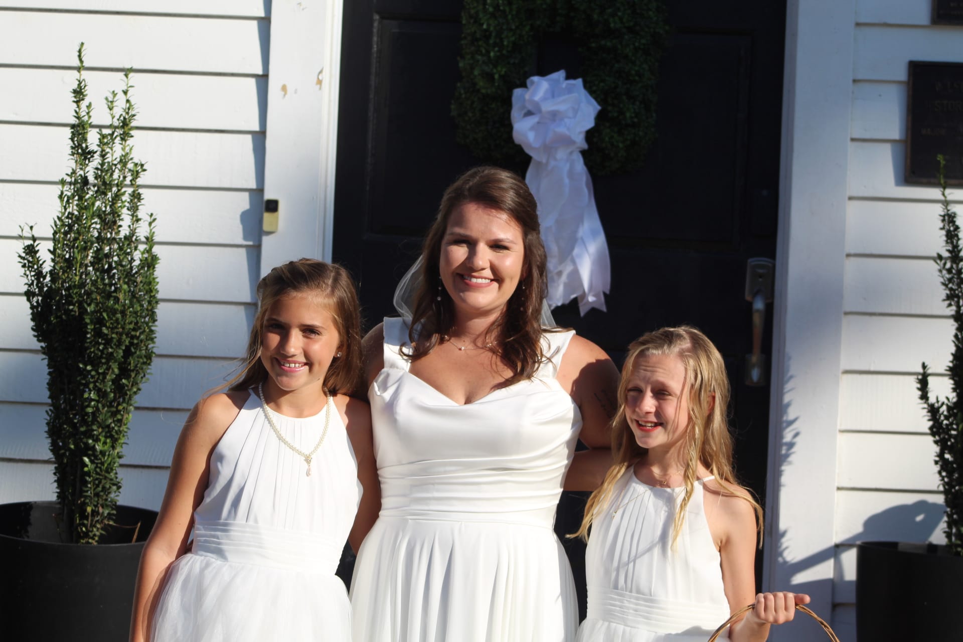 A bride in a flowing white gown smiles warmly with two young flower girls in white dresses on the front steps of Scarborough House, framed by a wreath-adorned black door with a white bow.