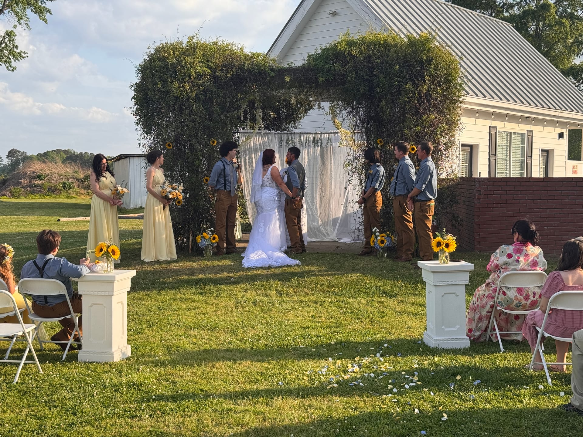 Bride and groom exchange vows beneath a lush greenery arch draped with white fabric and sunflowers on the ceremony lawn at Scarborough House historic estate venue.