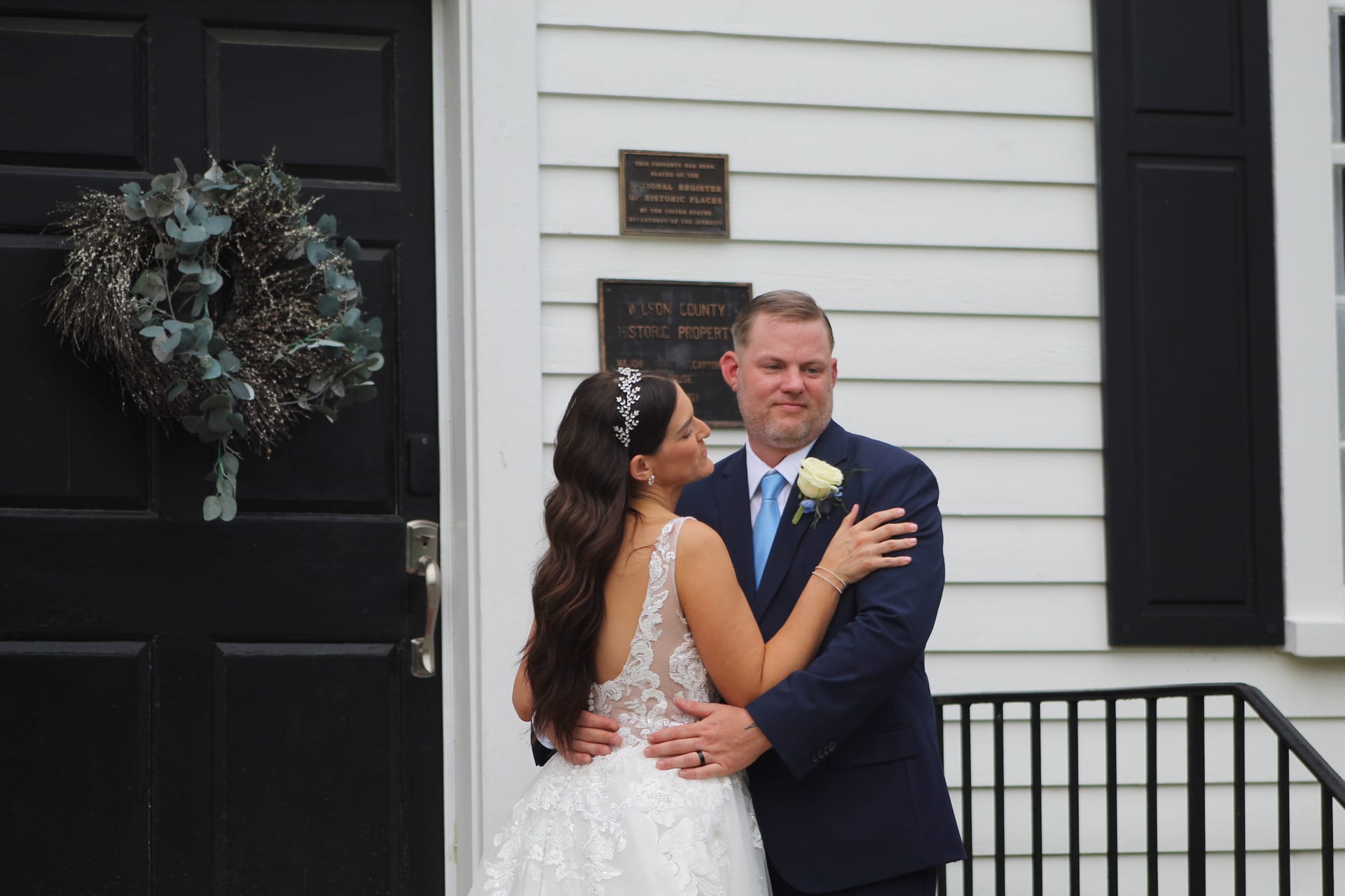 Bride in lace gown embracing groom in navy suit on the front steps of the historic 1821 Scarborough House manor, with a eucalyptus wreath and Wilson County Historic Property plaque visible on the white clapboard exterior.