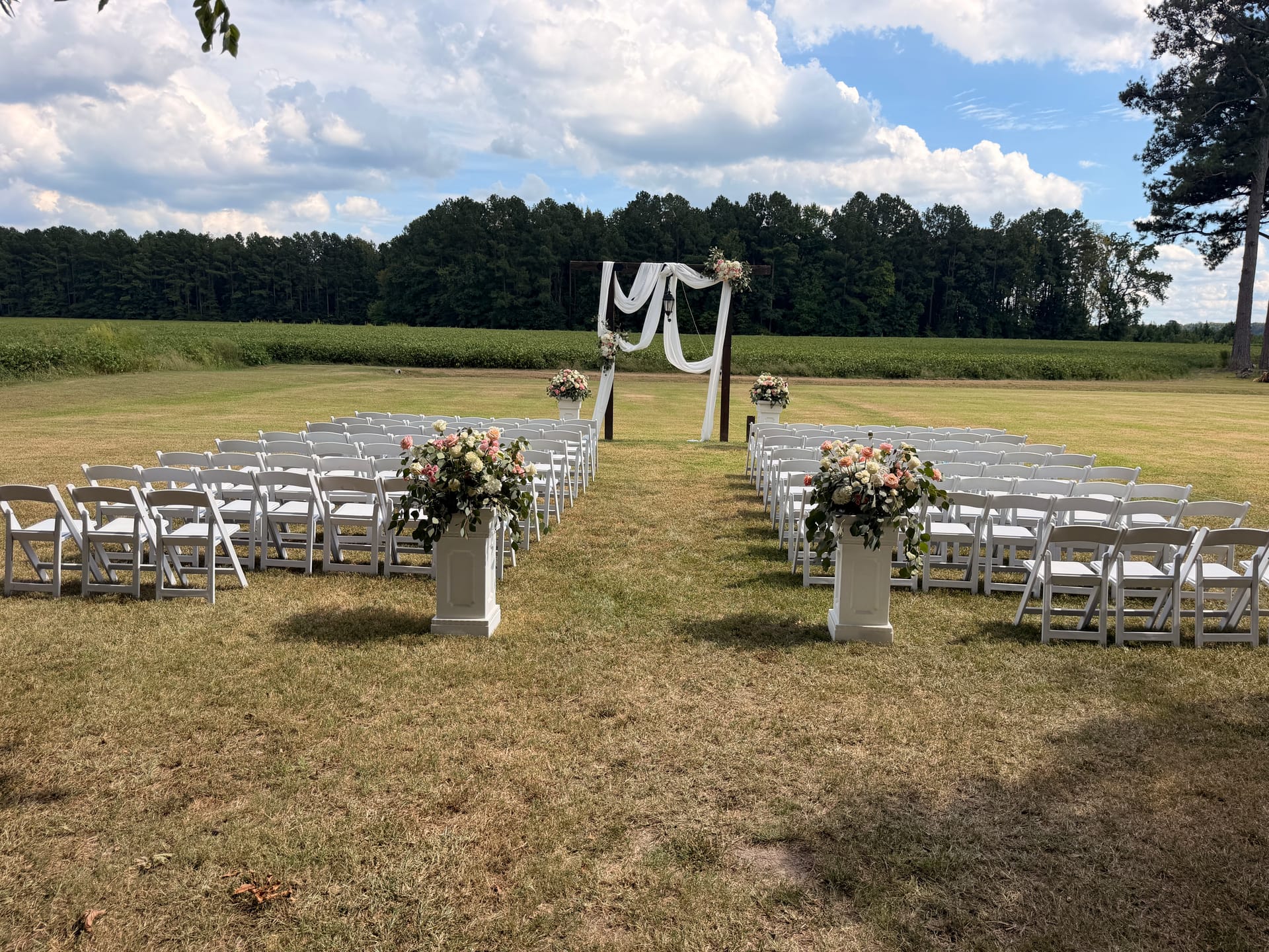 Outdoor wedding ceremony setup at Scarborough House featuring rows of white folding chairs, floral-topped pedestals lining the aisle, and a white draped arch adorned with pink and ivory blooms set against open farmland and a treeline.