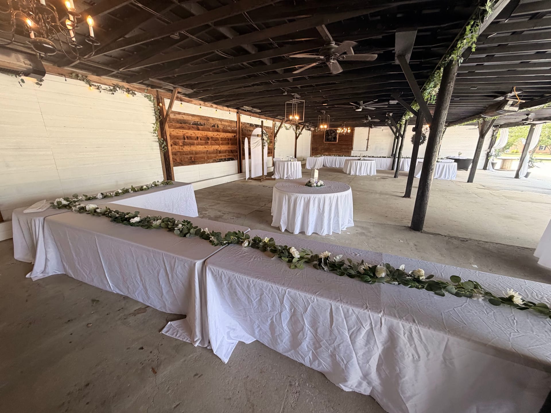 White linen-draped banquet tables adorned with green and white floral garlands fill the rustic magnolia pavilion at Scarborough House, set for a wedding reception.
