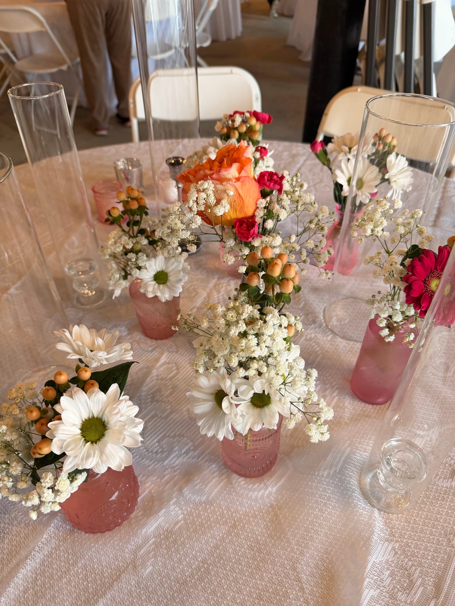 Pink glass bud vases filled with white daisies, baby's breath, orange roses, and hypericum berries arranged as reception table centerpieces at Scarborough House wedding venue.