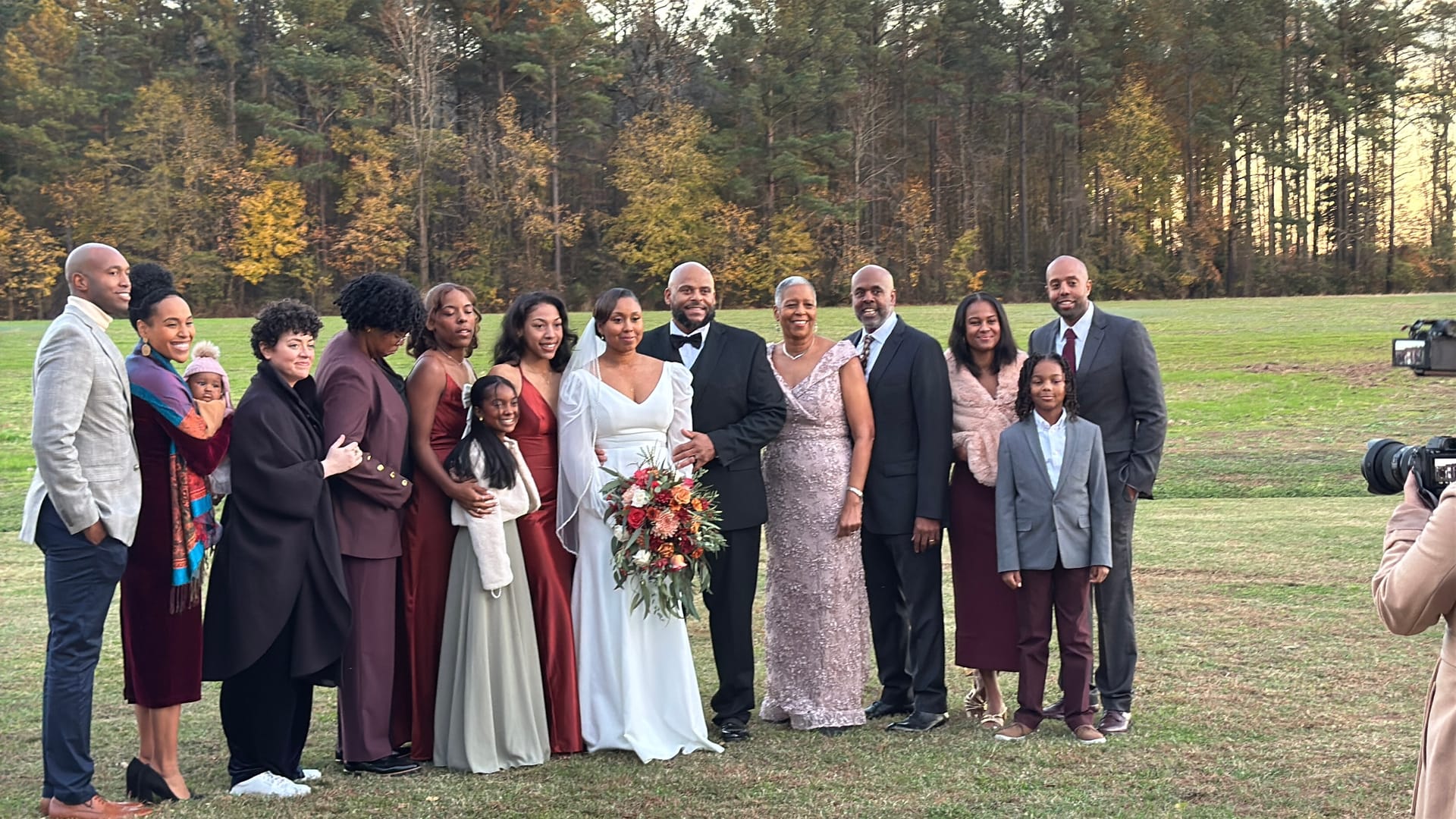 Bride and groom pose with a large family group on the open ceremony lawn at Scarborough House, surrounded by autumn foliage and pine trees in the background.