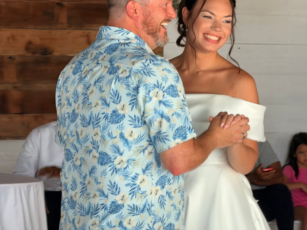 A beaming bride in an off-shoulder white gown shares a joyful first dance with a man in a floral shirt inside the rustic grand hall at Scarborough House.