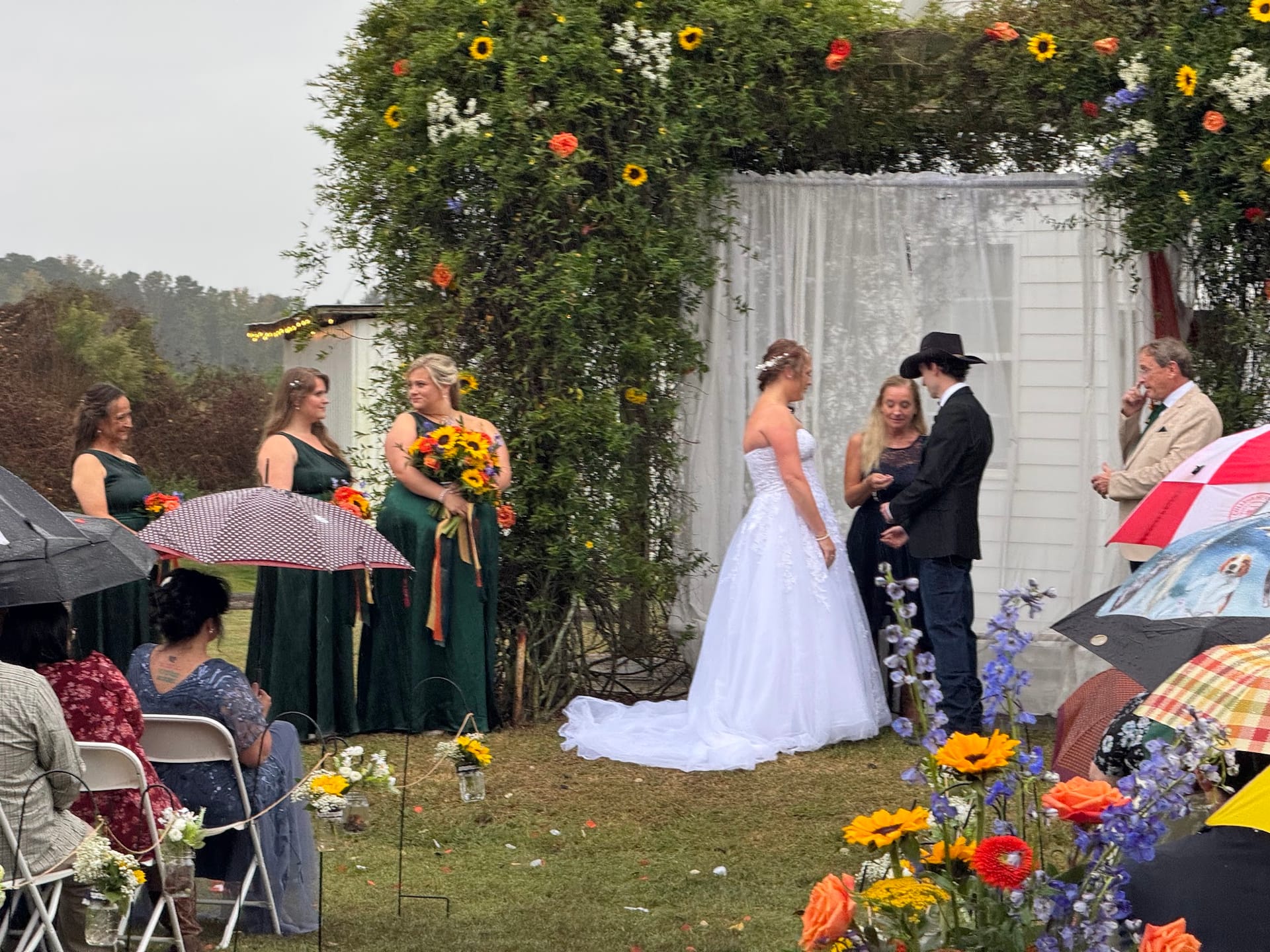 Bride and groom exchange vows beneath a lush floral arch adorned with sunflowers on the ceremony lawn at Scarborough House, surrounded by bridesmaids in emerald green and seated guests holding colorful umbrellas.