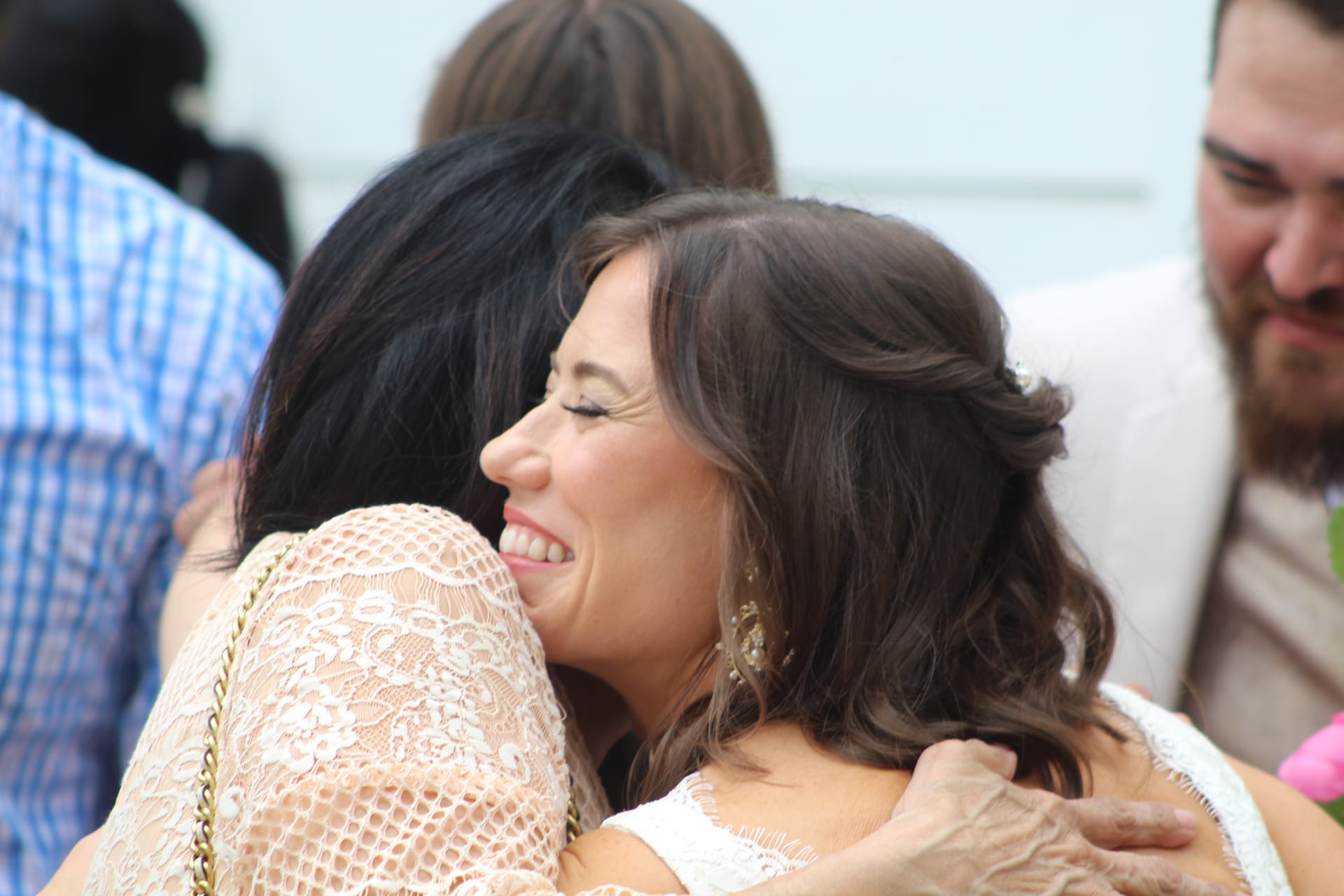 A smiling bride in a white dress shares a heartfelt hug with a guest in a lace top during an outdoor wedding celebration.