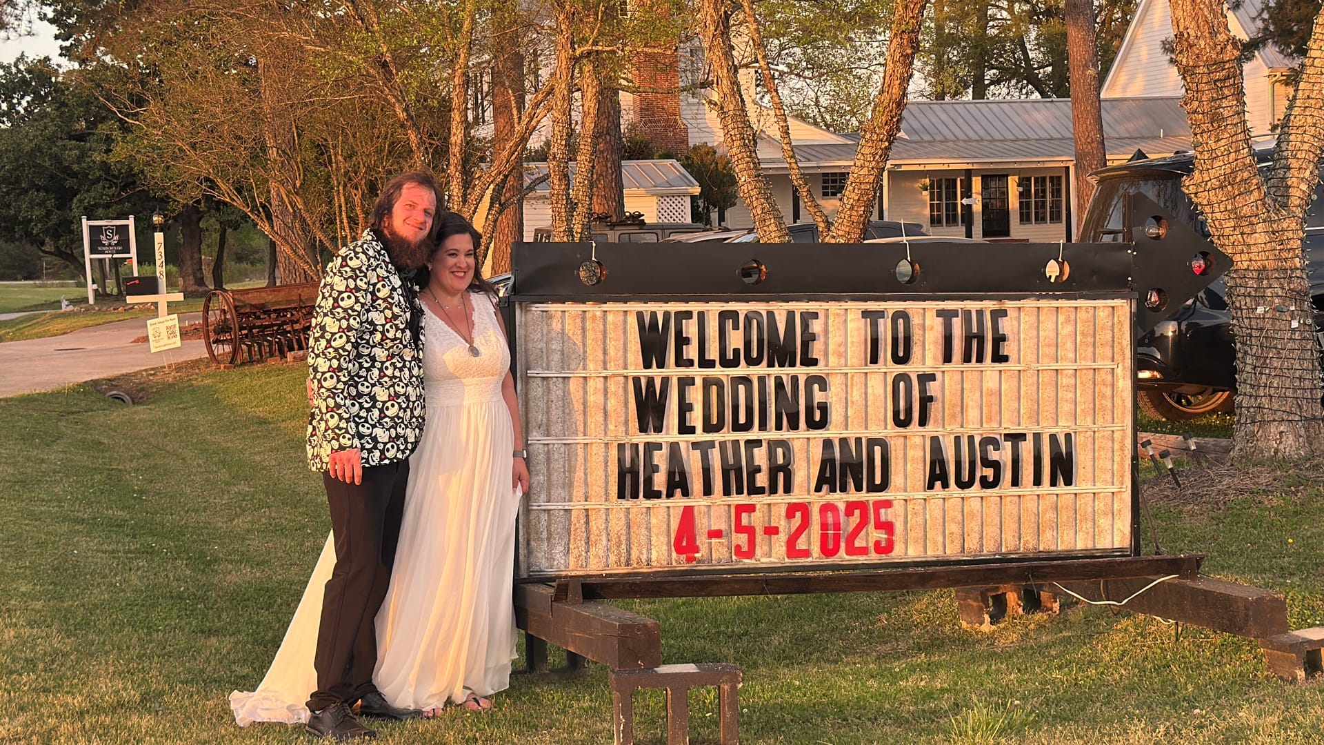 Bride and groom smiling together beside a lit marquee welcome sign at Scarborough House on their wedding day, April 5 2025, surrounded by mature trees in warm golden-hour light.