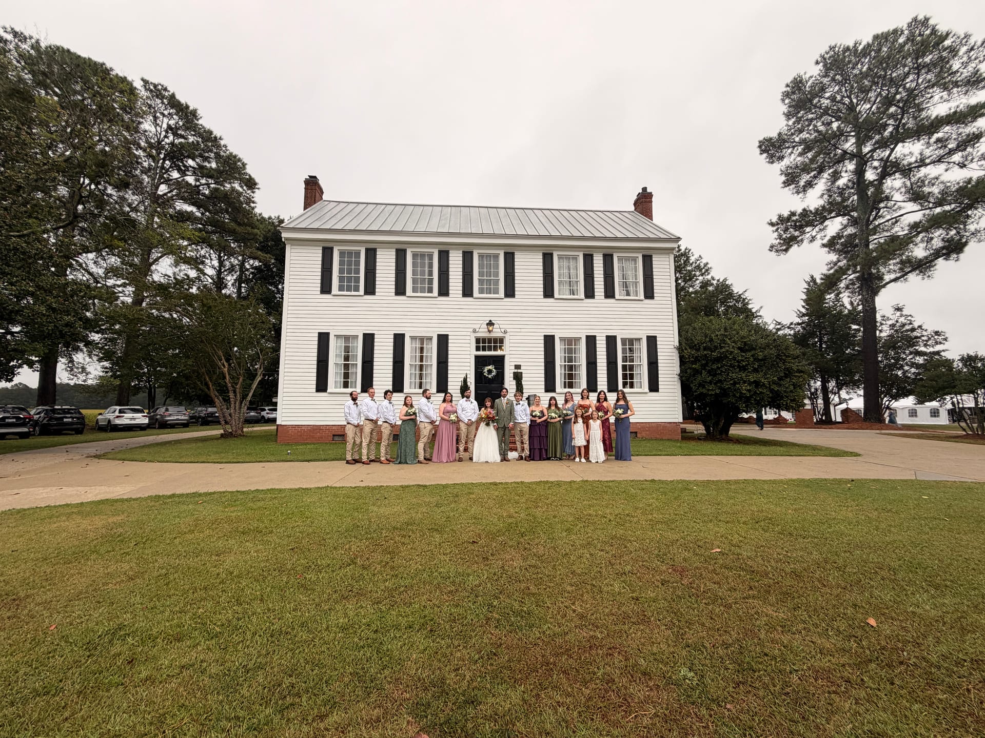 Full wedding party posed in front of the white 1821 Scarborough House manor with black shutters and brick chimneys on an overcast autumn day.