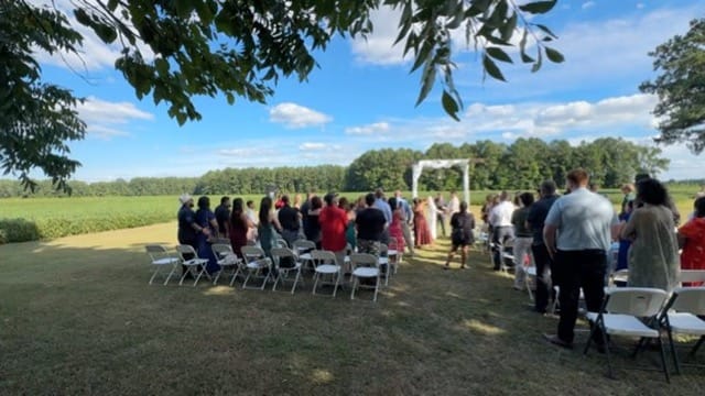 Outdoor wedding ceremony on the historic Scarborough House estate lawn with guests gathered before a white arch framed by open farmland fields and a tree-lined horizon under a bright blue sky.
