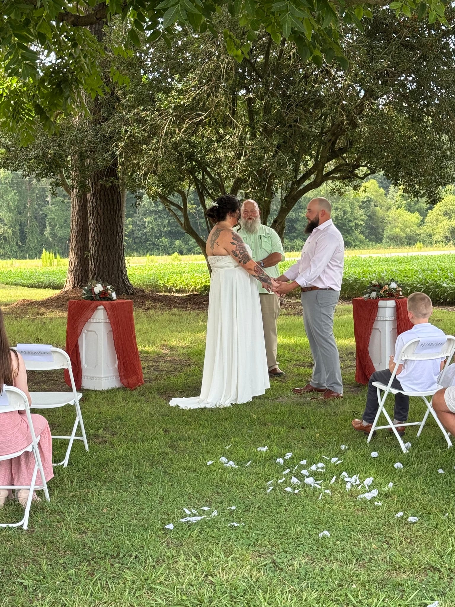 A bride and groom hold hands during an intimate outdoor wedding ceremony beneath sprawling oak trees at Scarborough House, with lush farmland fields stretching into the background.