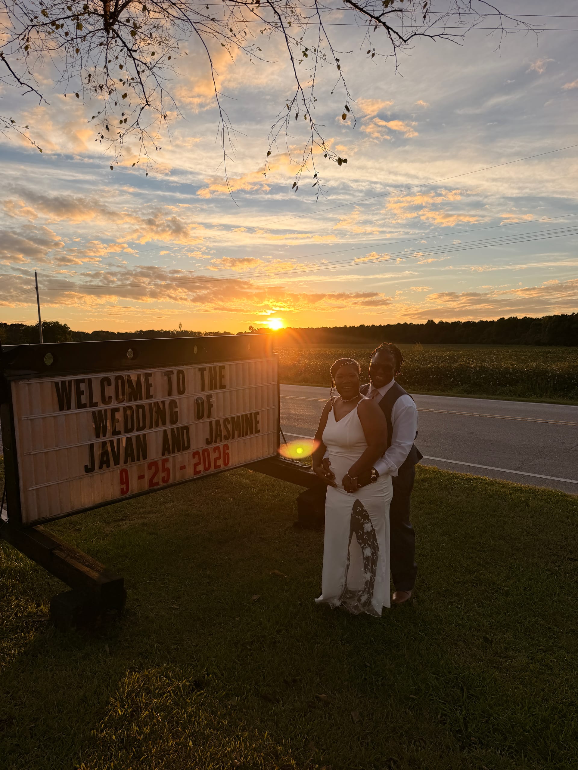 Bride and groom embrace at sunset beside a marquee sign welcoming guests to the wedding of Javan and Jasmine at a rustic countryside venue with a glowing orange sky.