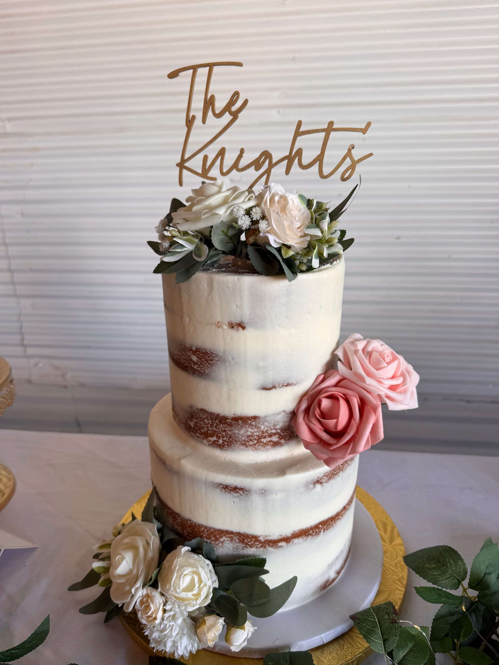 Two-tier semi-naked wedding cake decorated with blush and ivory roses and a gold laser-cut The Knights cake topper displayed on a gold charger plate at Scarborough House.