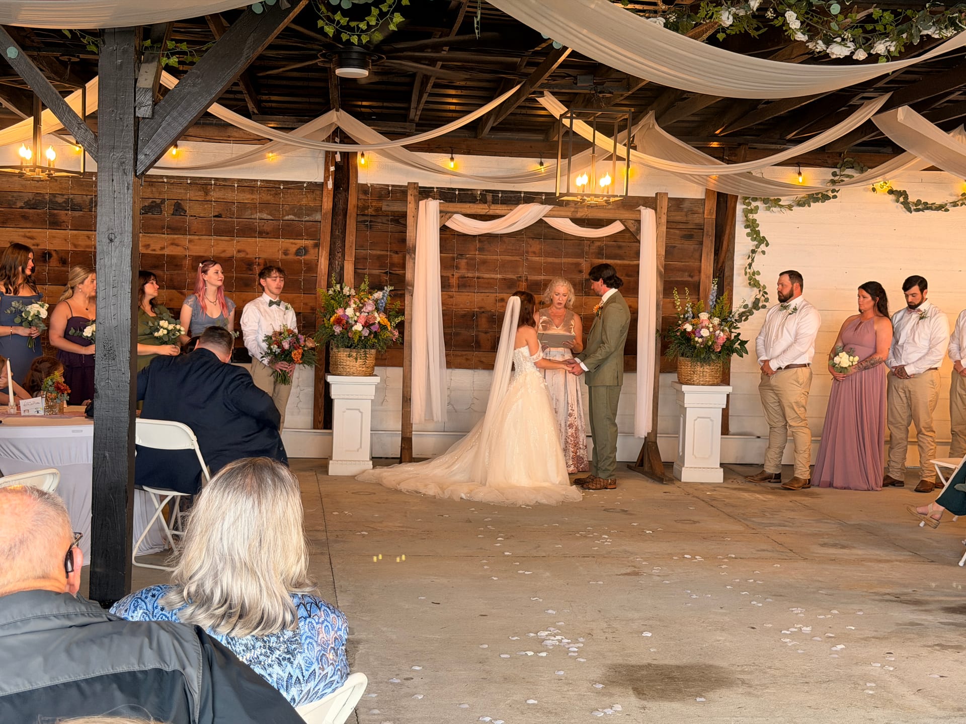 Bride and groom exchange vows beneath a draped fabric arch adorned with florals and string lights inside a rustic barn-style venue, surrounded by their wedding party and seated guests.