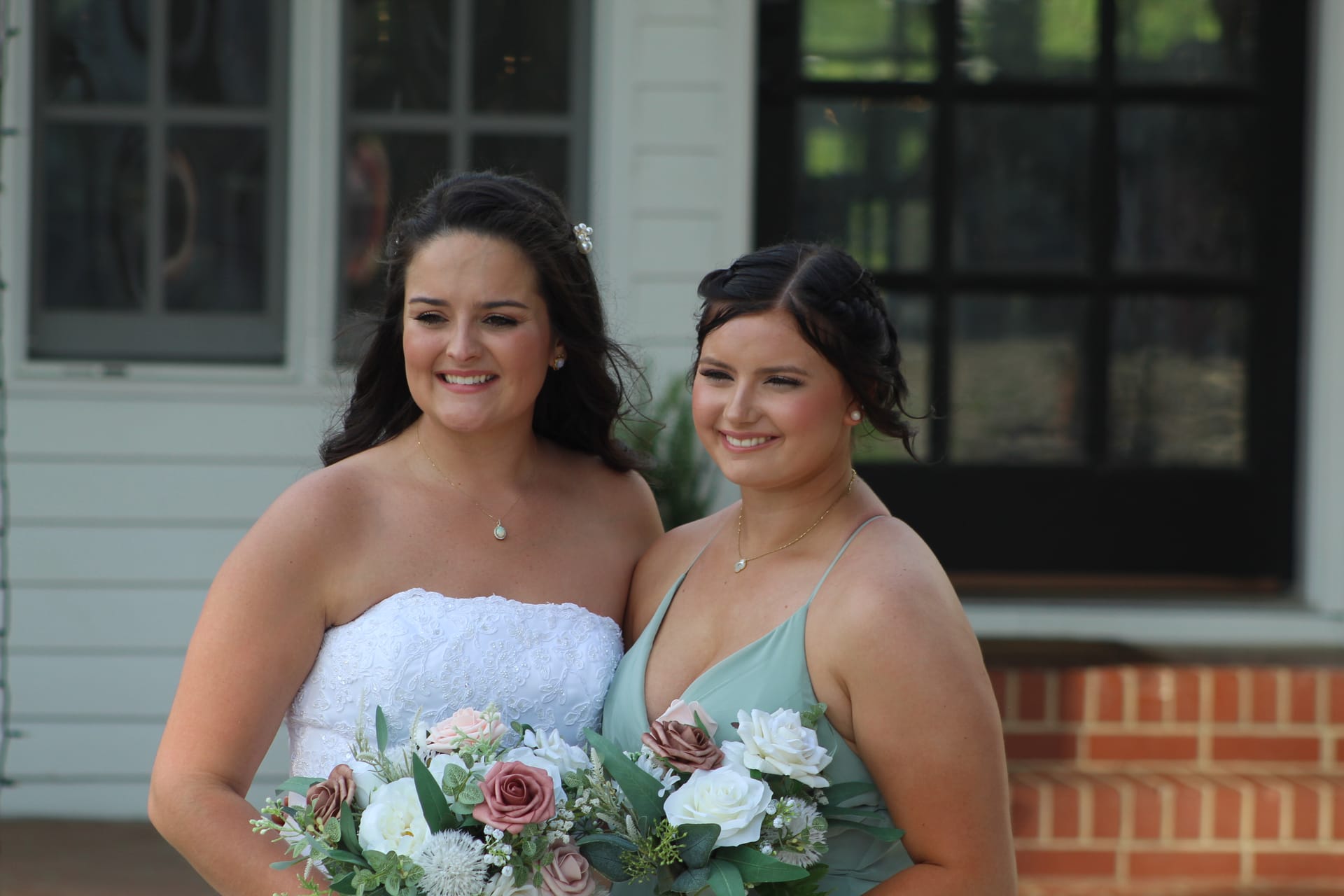 Bride in white strapless lace gown and bridesmaid in sage green dress smile together holding rose bouquets in front of the historic 1821 Scarborough House manor facade.