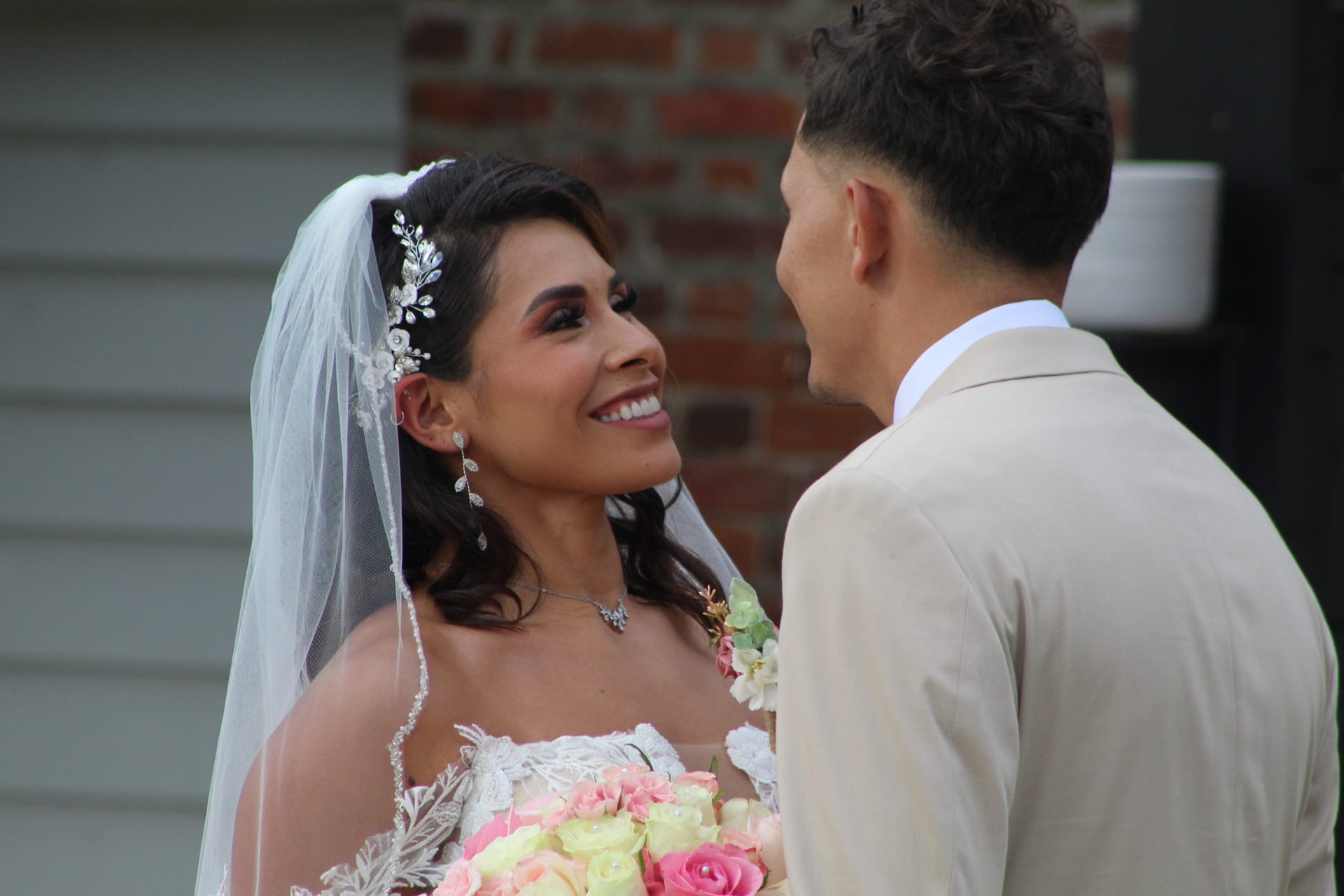 A glowing bride in a lace gown and floral veil smiles warmly up at her groom in a tan suit during their first look at Scarborough House historic estate.
