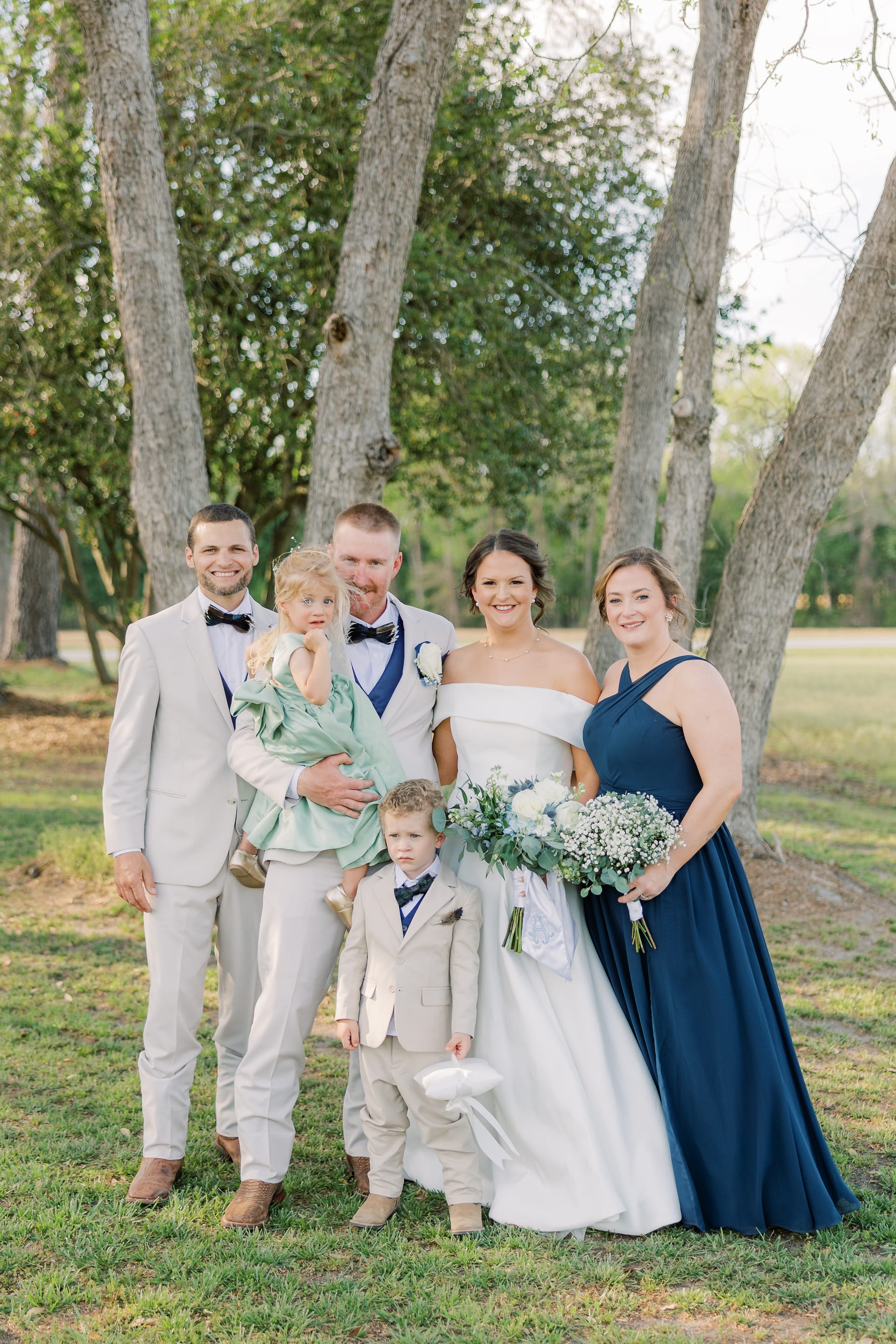 Wedding party group portrait with bride in off-shoulder gown, groom, groomsman, bridesmaid in navy dress, flower girl, and ring bearer standing among oak trees at Scarborough House.