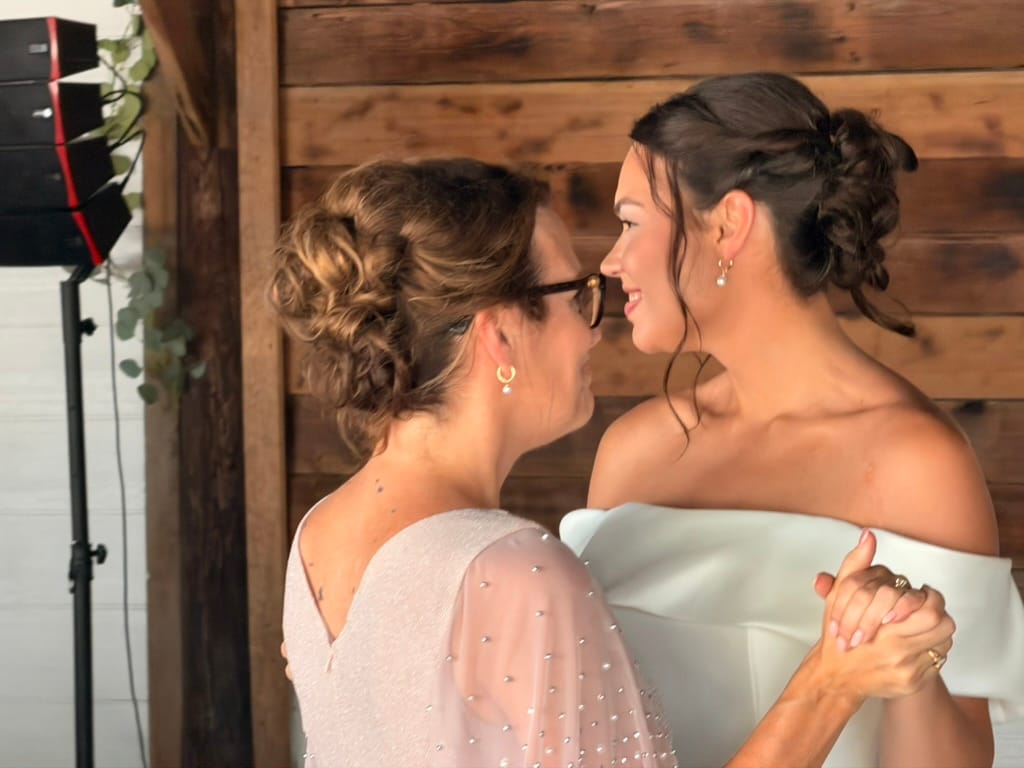 Bride in an off-shoulder white gown shares a tender face-to-face moment with a guest in a blush embellished dress against a rustic wood-paneled wall at Scarborough House.