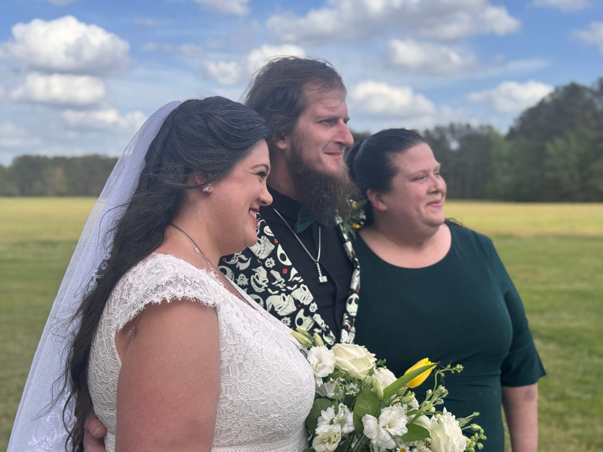 Smiling bride in white lace gown holding a bouquet of white flowers and yellow tulips stands with her bearded groom and a guest on the open farmland fields at Scarborough House under a partly cloudy blue sky.