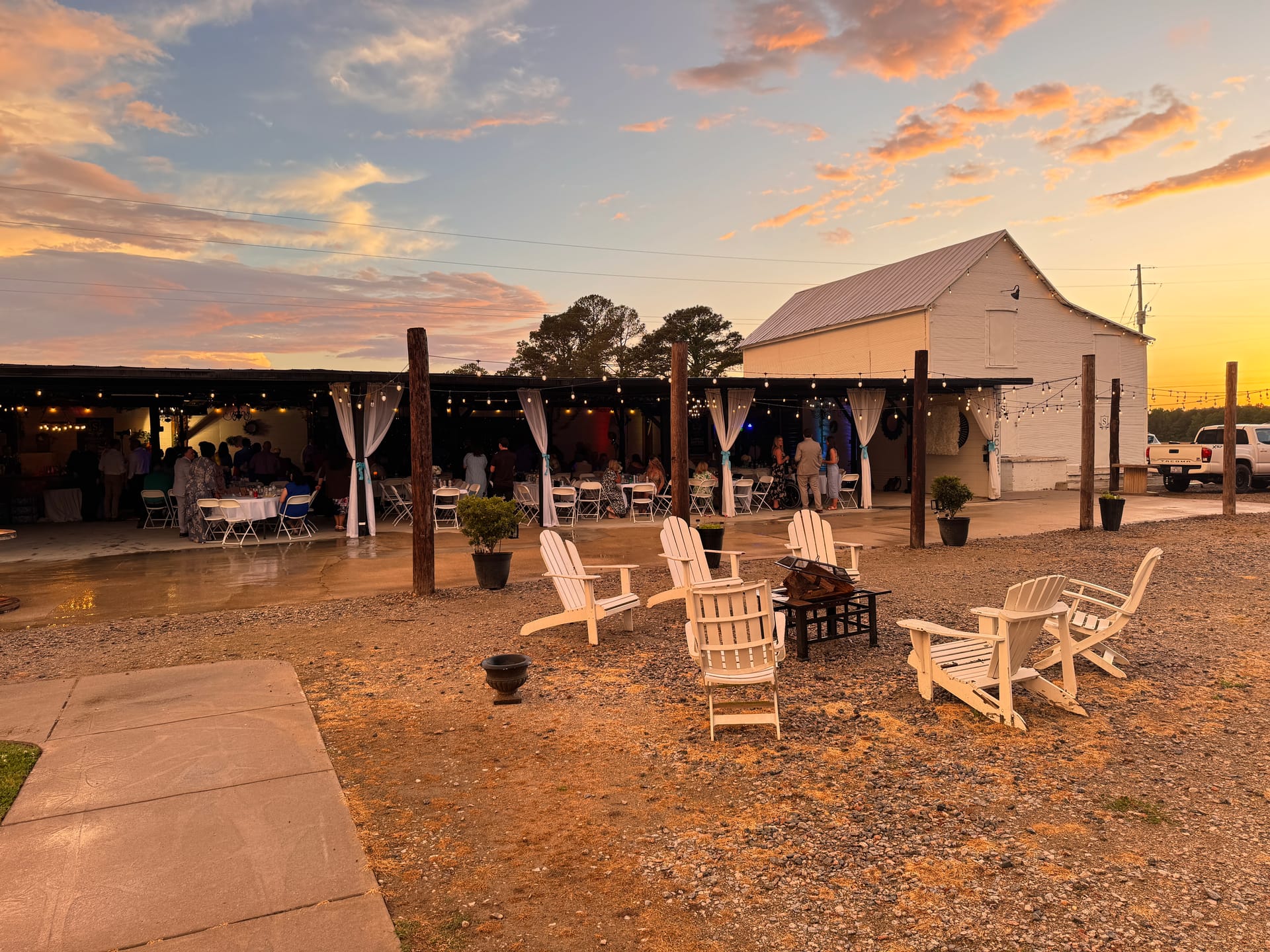 White Adirondack chairs arranged around a fire pit at dusk with string lights glowing over a lively outdoor wedding reception beneath a rustic covered pavilion at sunset.