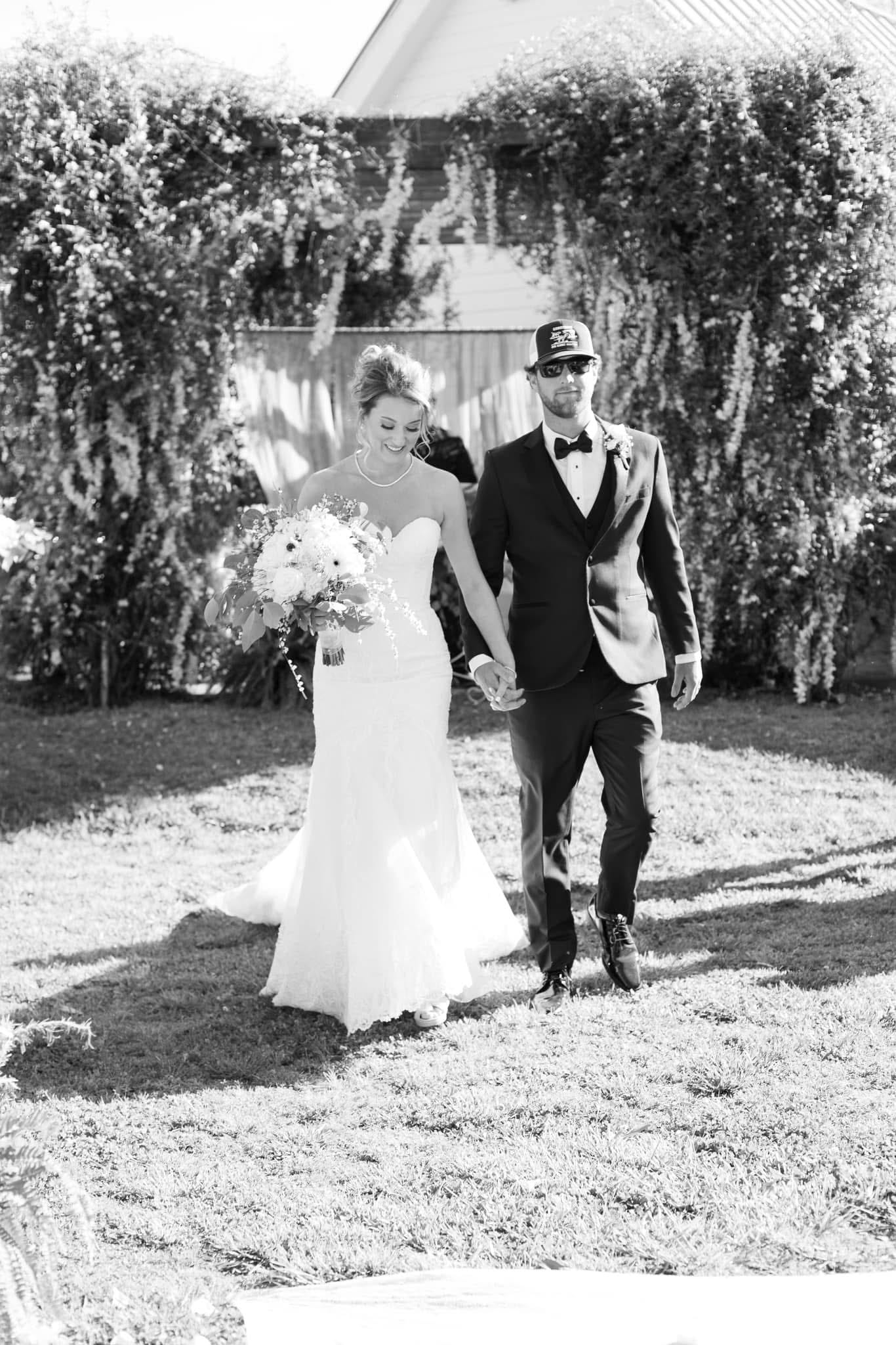 Bride in a fitted lace gown holding a large floral bouquet walks hand-in-hand with the groom in a dark suit and bow tie across the sunlit ceremony lawn beneath a lush ivy-covered arch at Scarborough House.